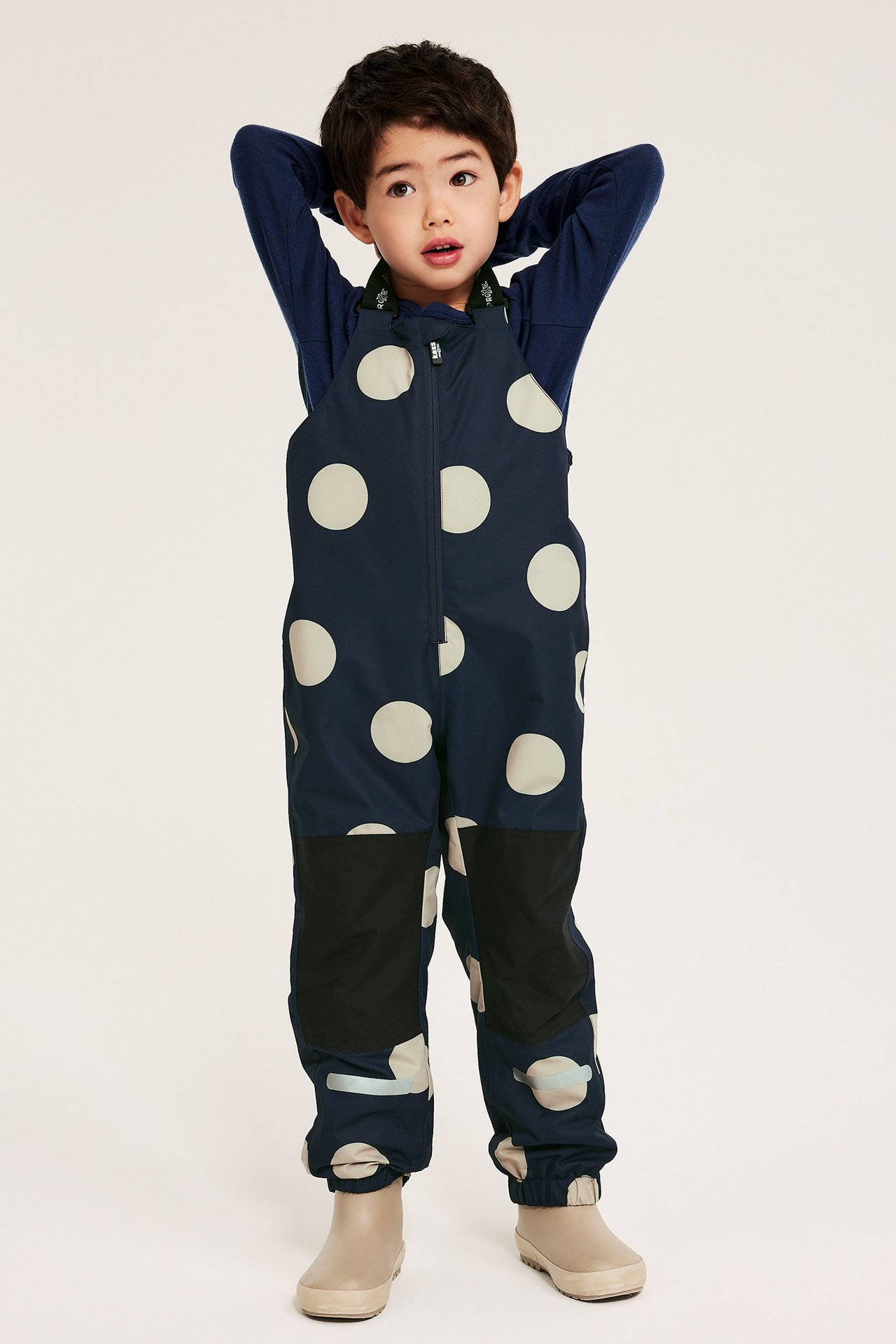 Frontvisning av et barn i mørkeblå prikkete skallbukser over en mørkeblå langermet genser og beige støvler.