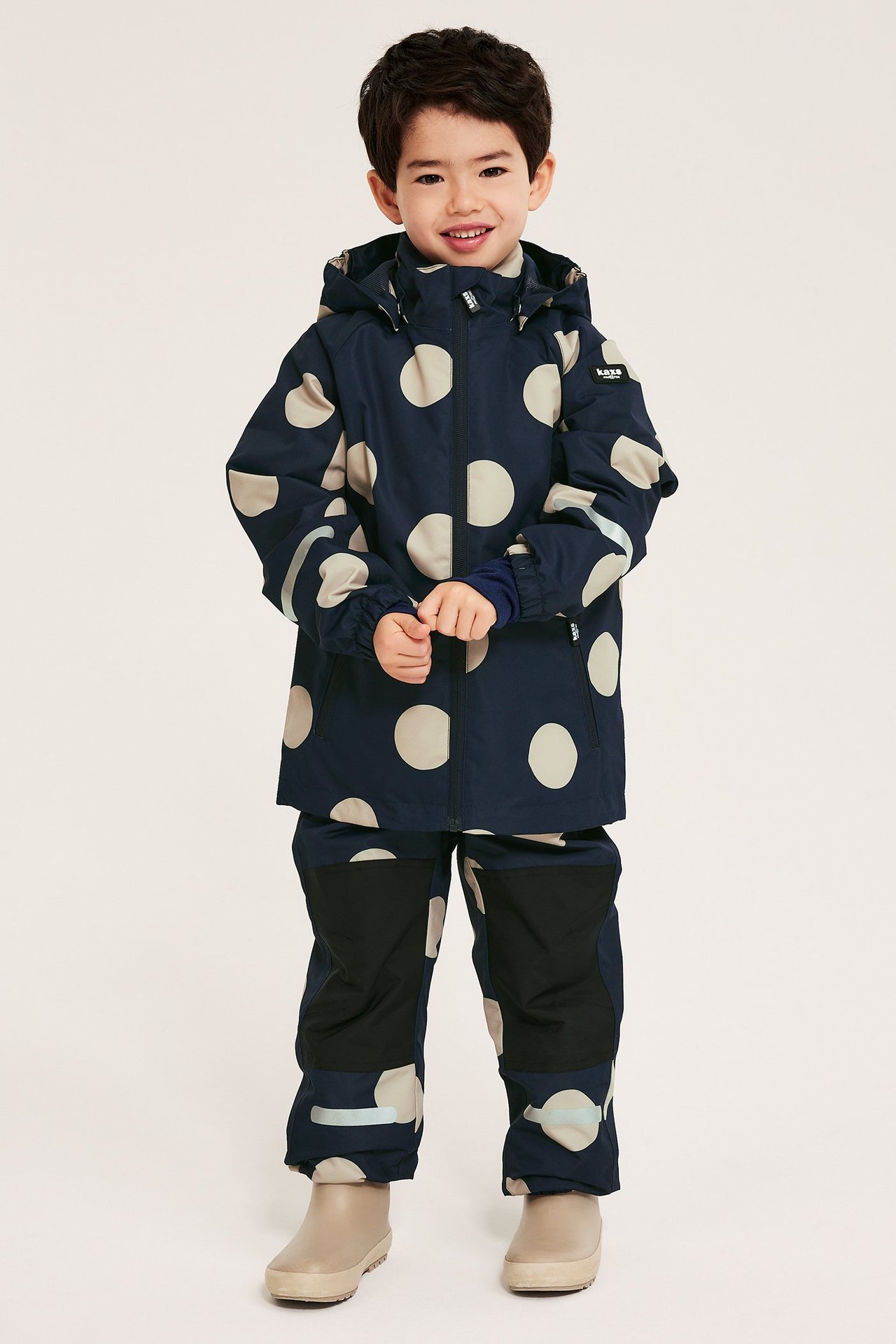 Edestä kuvattuna lapsi tummansinisessä vedenpitävässä kuoritakissa, jossa beigenvärisiä pilkkuja, ja yhteensopivissa housuissa.