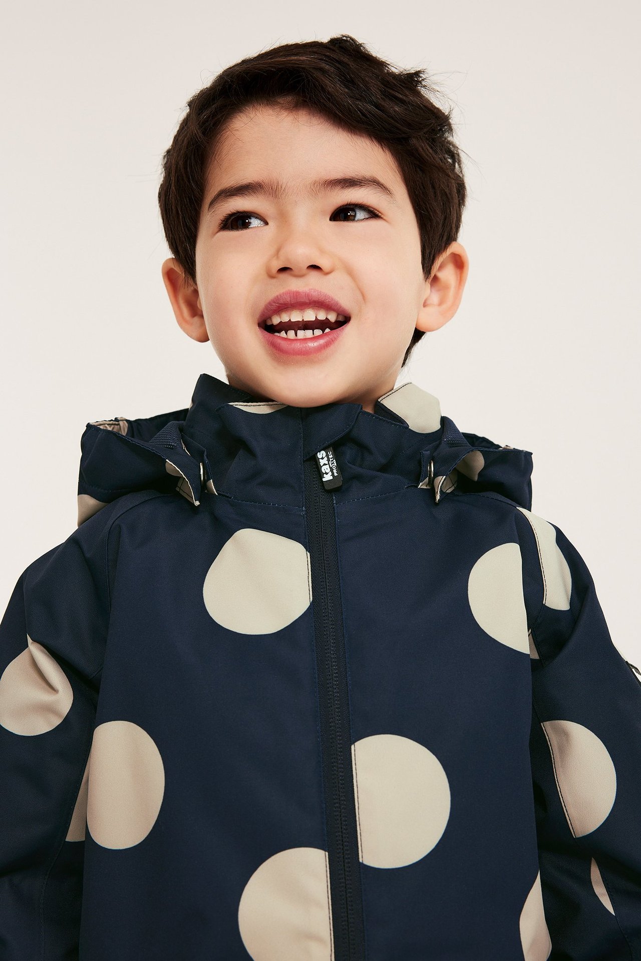 Framifrån: En glad pojke i en mörkblå skaljacka med beigea prickar, huva och dragkedja fram.