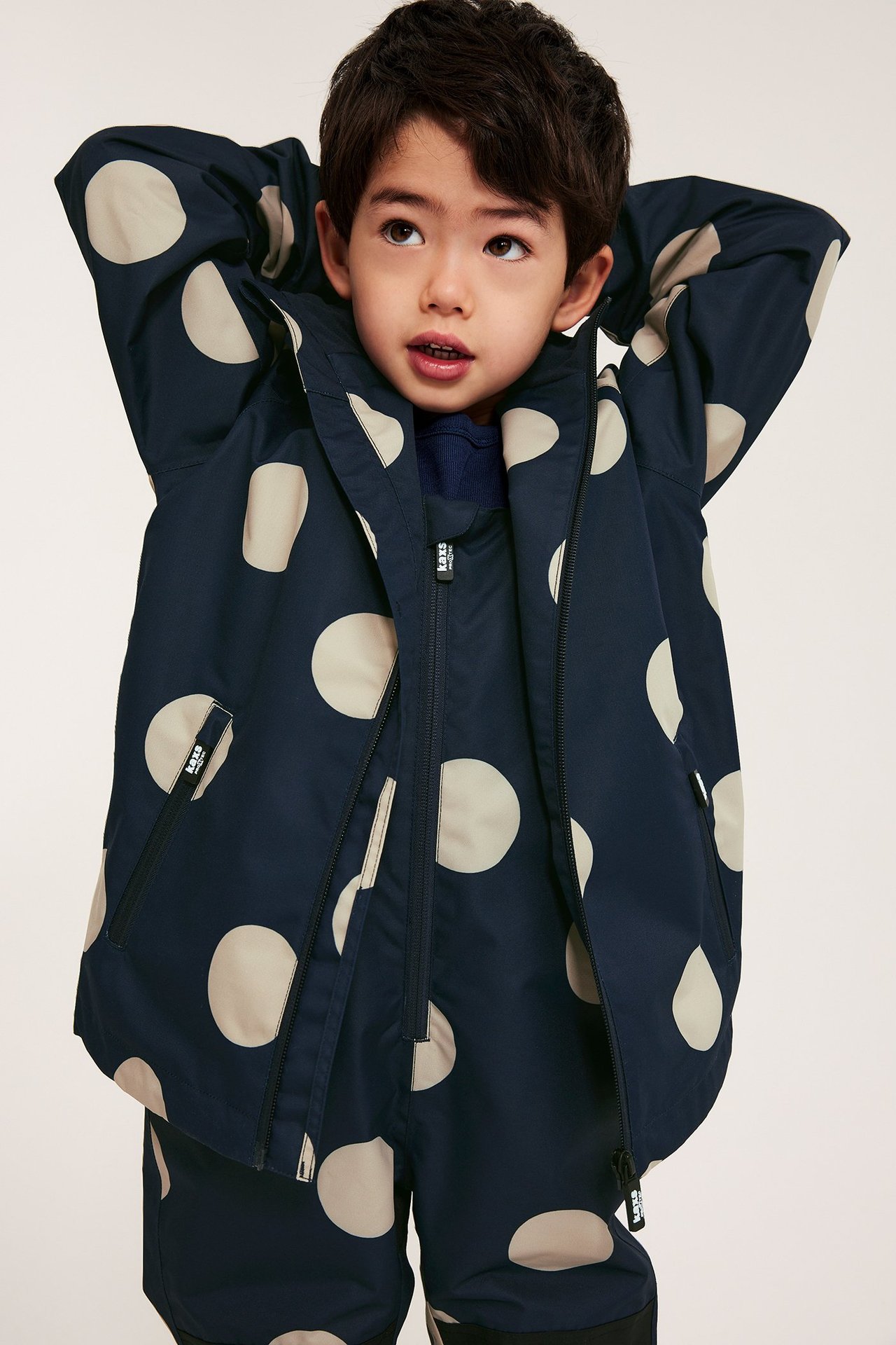 Framifrån: Ett barn i mörkblå, beigeprickig skaljacka, delvis öppen, och matchande skalbyxor.