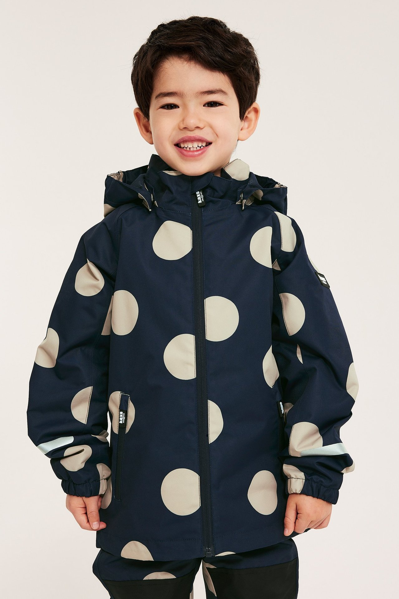 Edestä: hymyilevä lapsi tummansinisessä beigepilkullisessa vedenpitävässä kuoritakissa ja housuissa.
