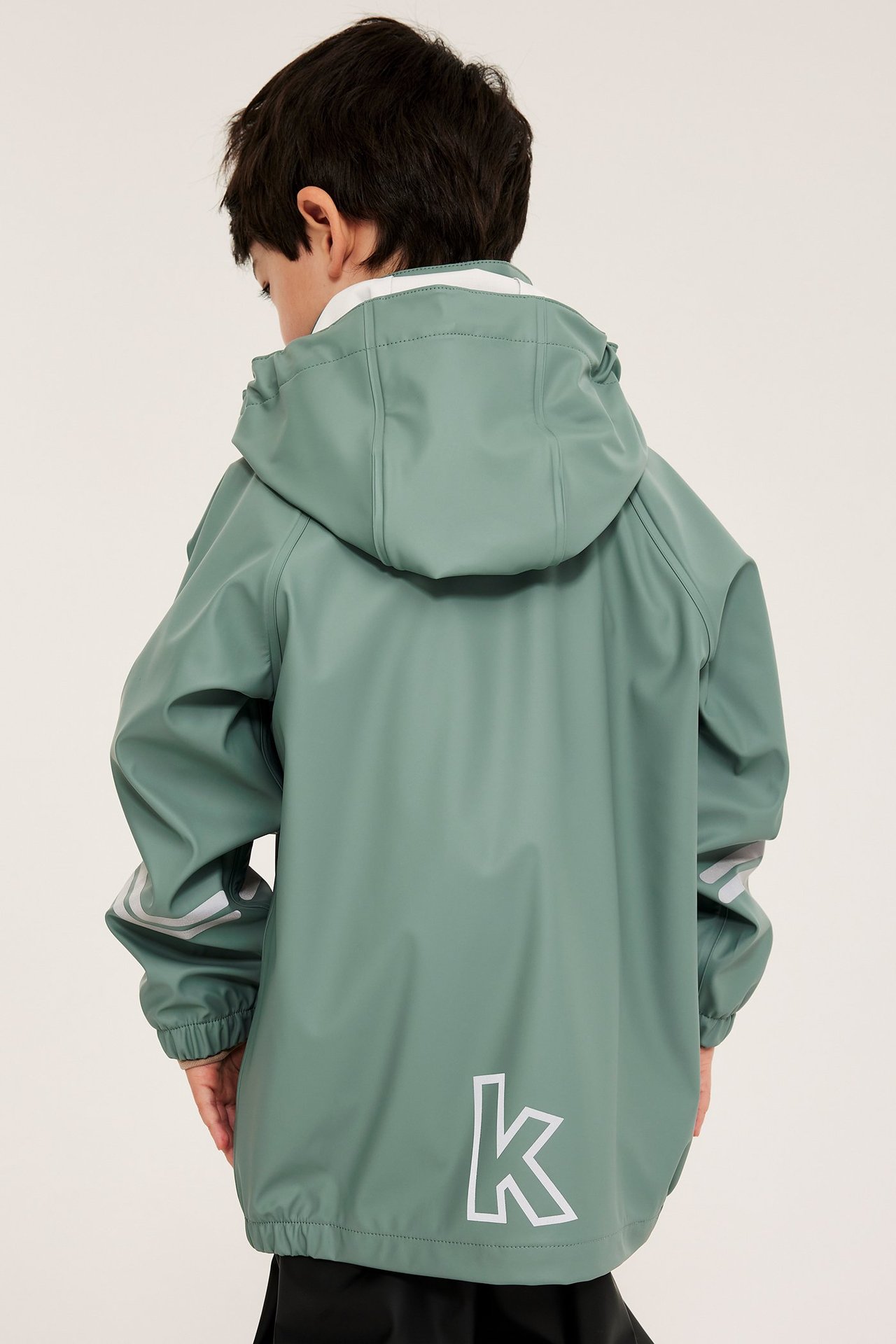 Ryggvy av ett barn i ljusgrön regnjacka med huva, reflexränder på ärmarna och ett 'k'-märke.