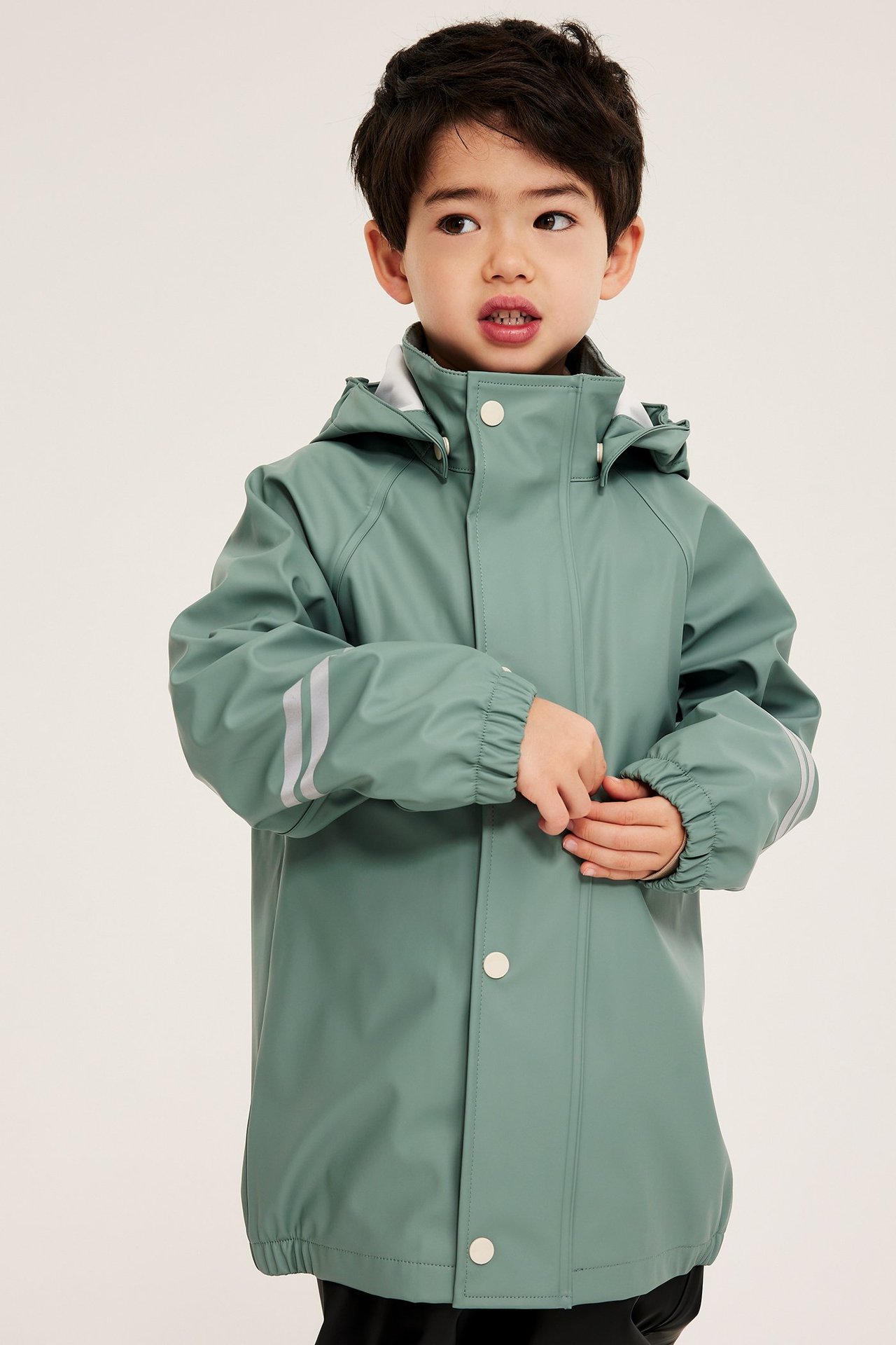 Framifrån, barn i ljusgrön regnjacka med huva, knappar och reflexränder.