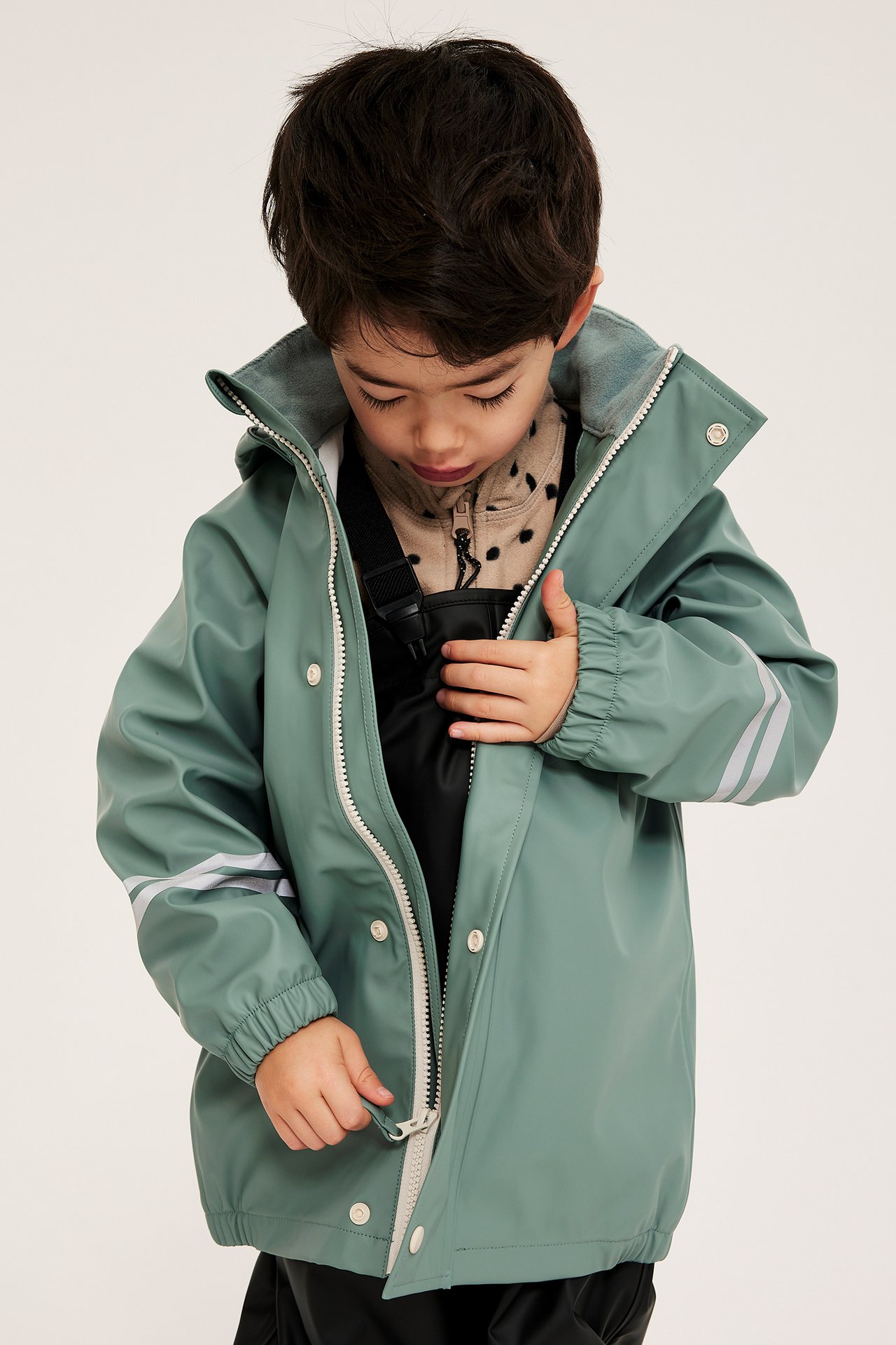 Främre vy av ett barn som drar upp en ljusgrön regnjacka med reflexränder över en beige prickig topp.