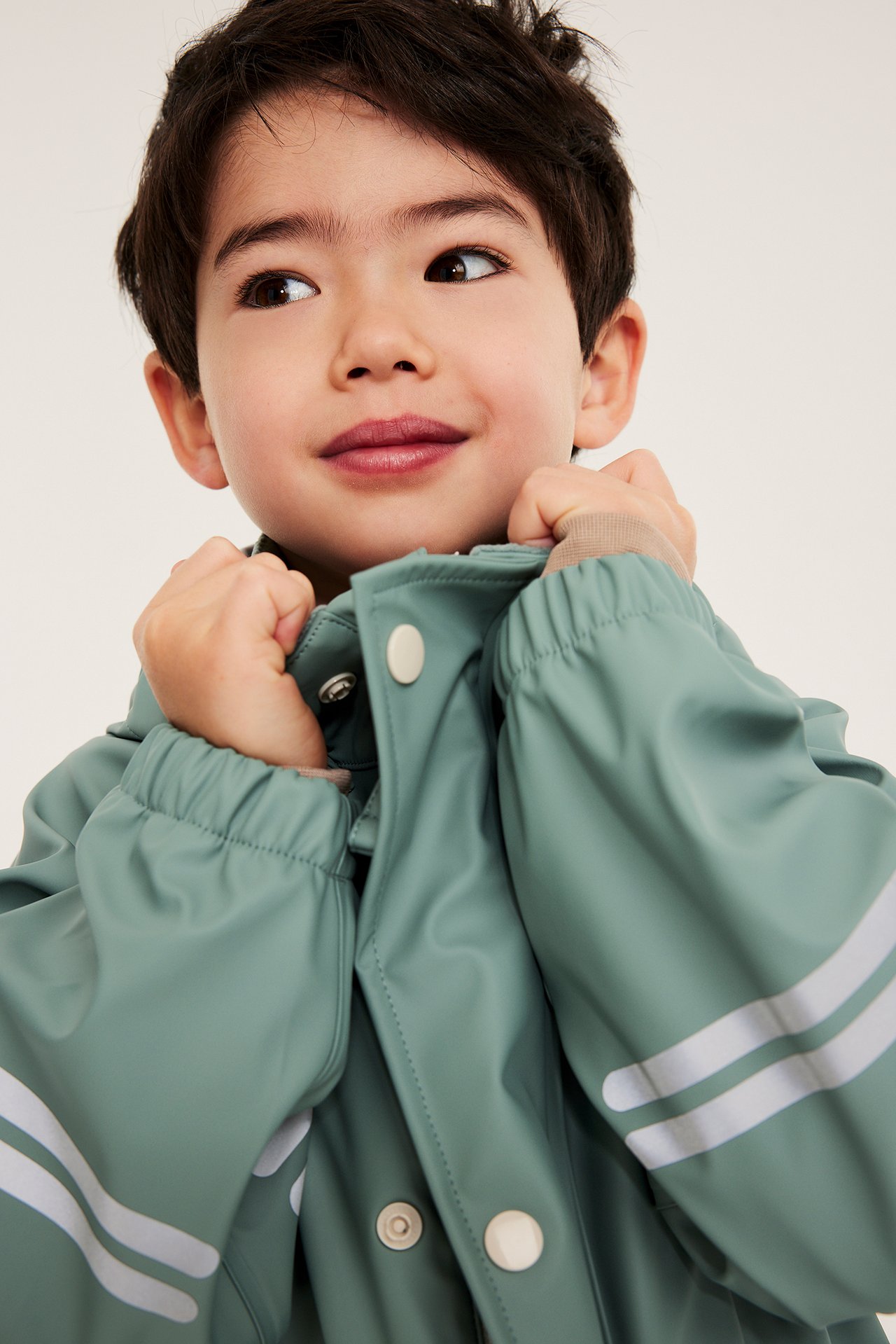 Närbild: Ett barn i en ljusgrön regnjacka med vita tryckknappar och reflexränder.