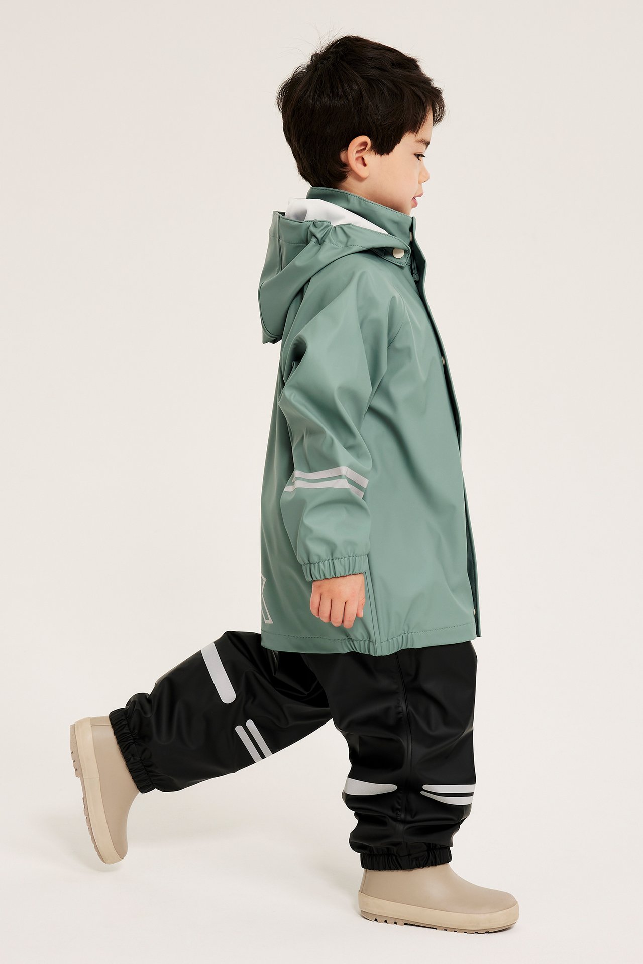 Sidovy av ett barn i ljusgrön regnjacka med huva och reflexer, svarta byxor, beiga stövlar.