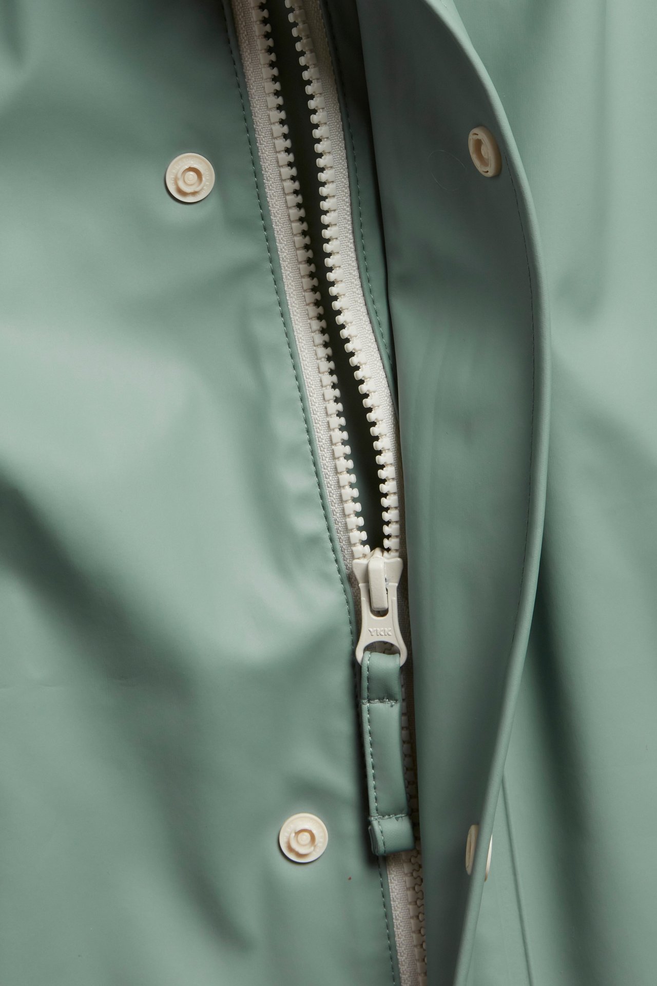 Zbliżenie na przód jasnozielonej kurtki przeciwdeszczowej z białym zamkiem błyskawicznym i jasnymi zatrzaskami.