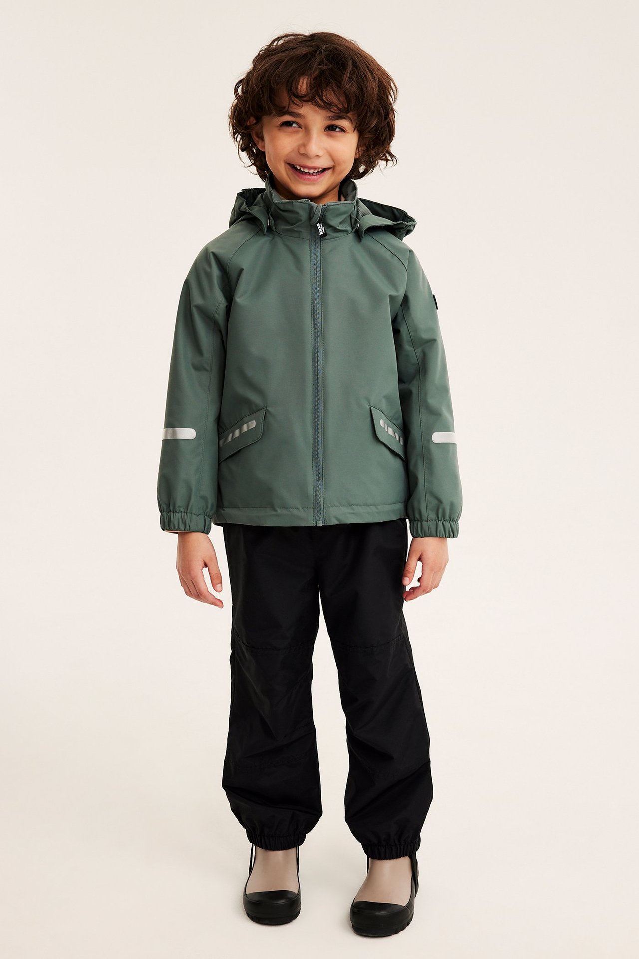 Edestäpäin hymyilevä lapsi yllään khakinvihreä vedenpitävä kuoritakki hupulla, mustat housut ja beiget kumisaappaat.