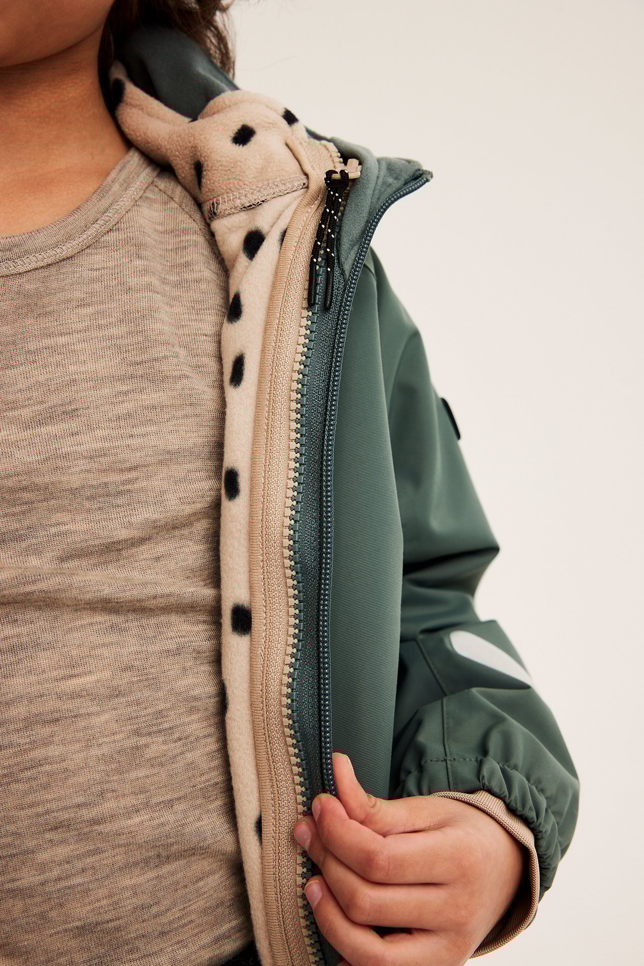 Lähikuva lapsesta avaamassa khakinvihreää kuoritakkia, joka paljastaa beigen pilkullisen fleecen ja vaaleanruskean t-paidan.