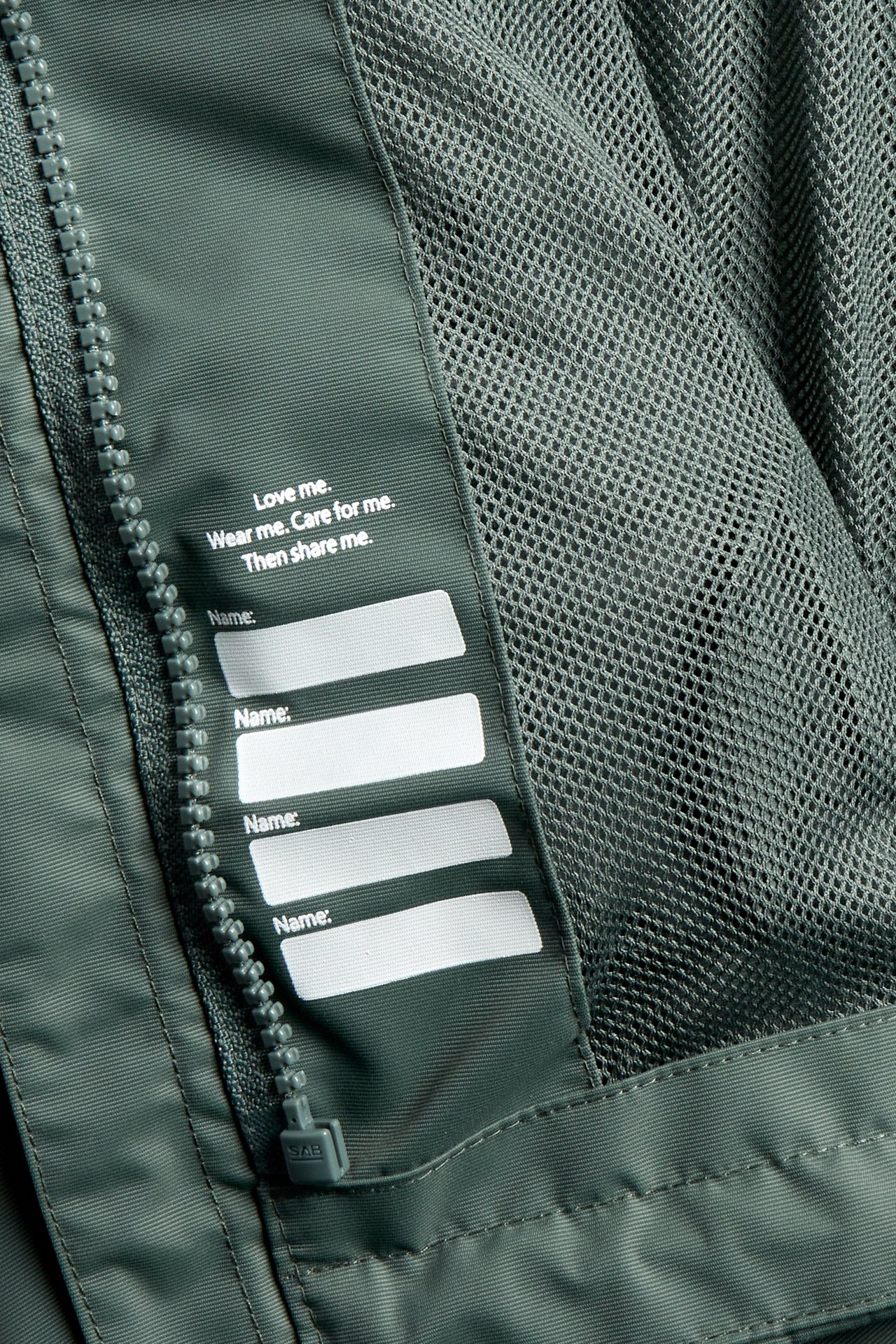 Närbild: Khakigrön vattentät skaljackas insida med dragkedja, meshfoder och namnetikett.