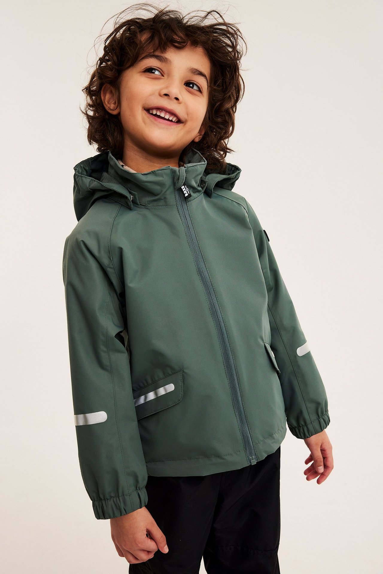 Edestäpäin hymyilevä lapsi yllään yksivärinen khakinvihreä kuoritakki vetoketjulla ja mustat housut.