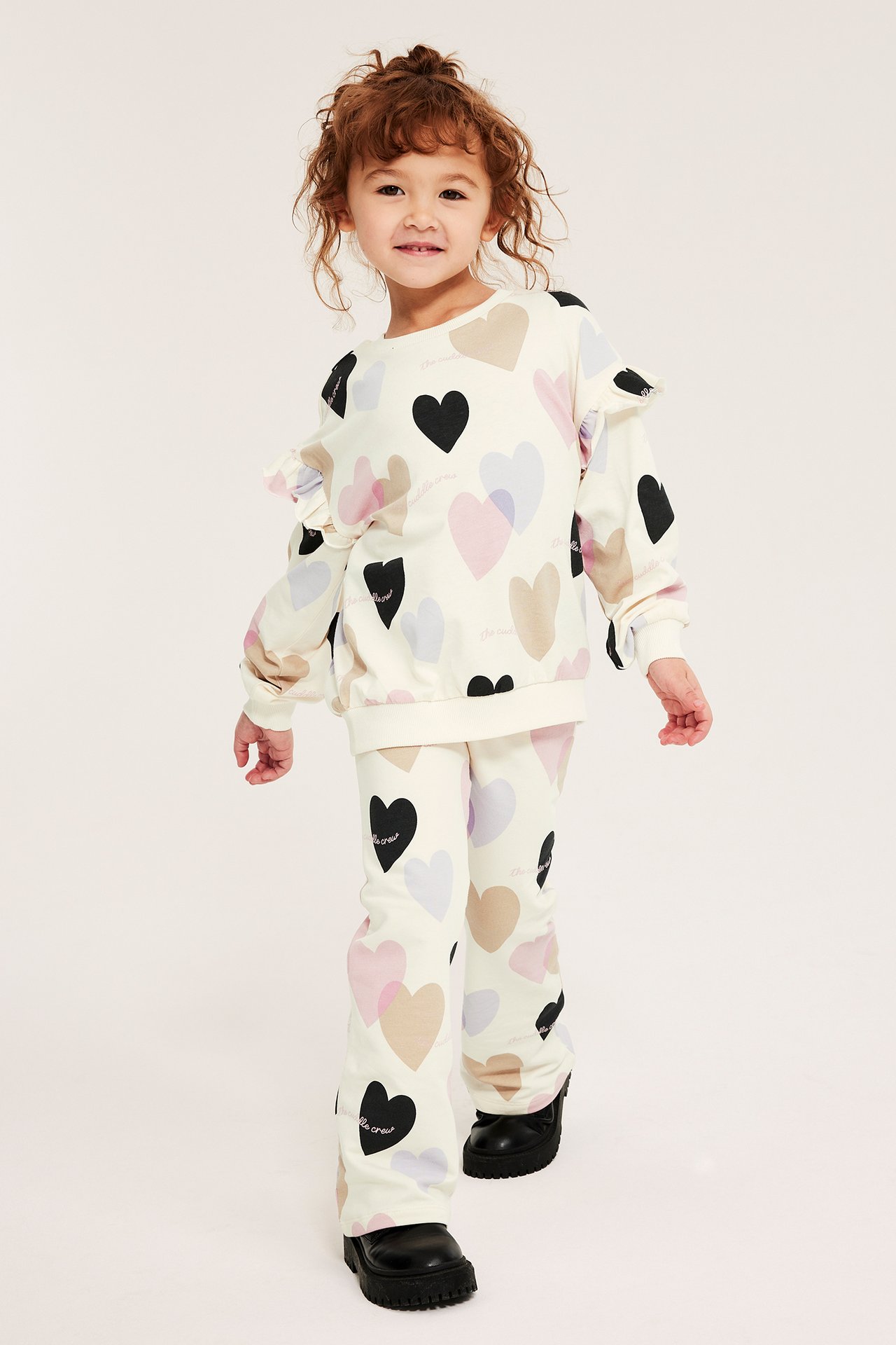 Widok z przodu dziecka w kremowej bluzie bawełnianej w serca, z falbankami i pasujących spodniach dzwonach.