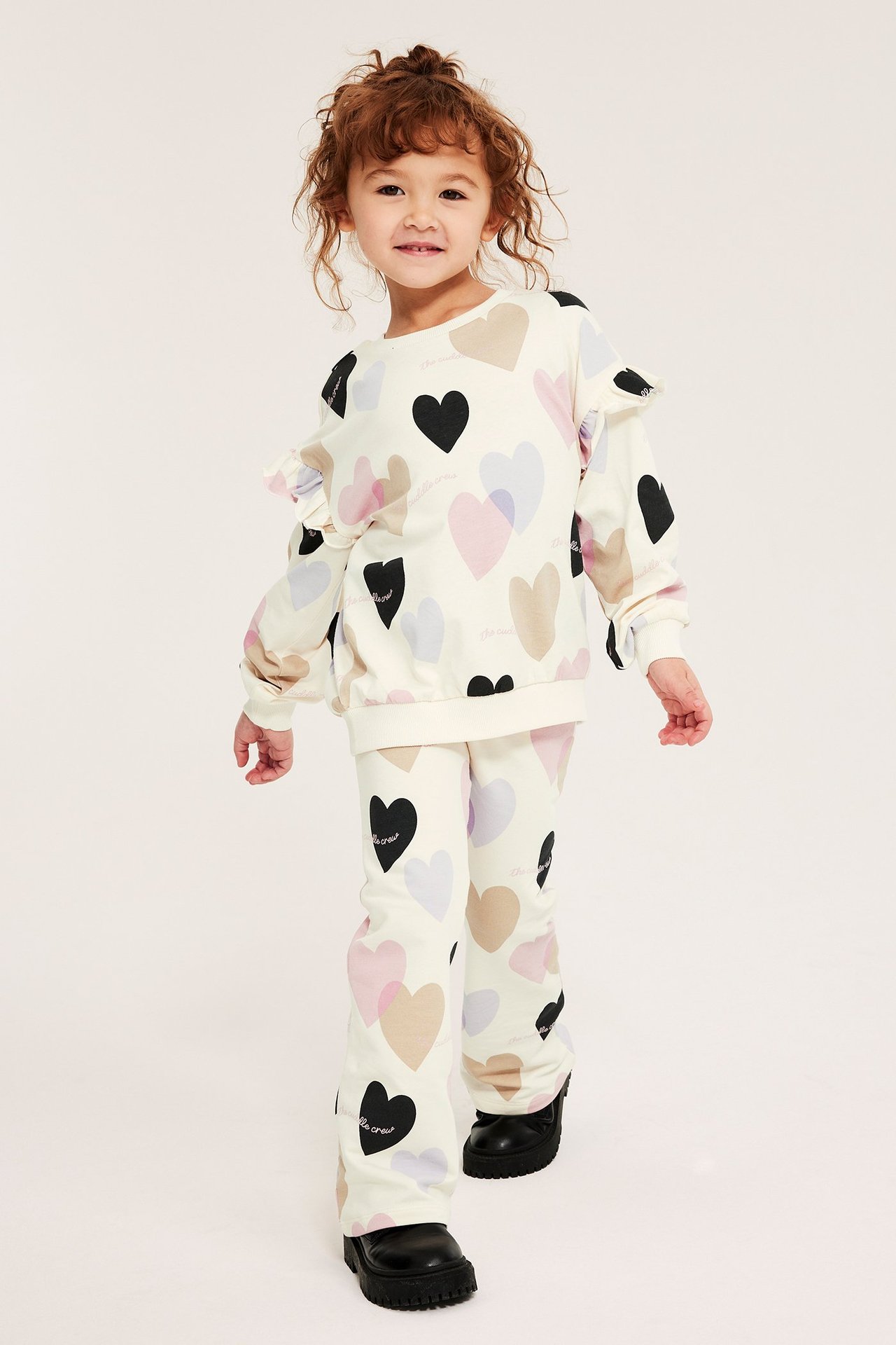 Frontbilde av et barn i en offwhite bomullssweatshirt med hjerteprint, rysjer og matchende utsvingte bukser.