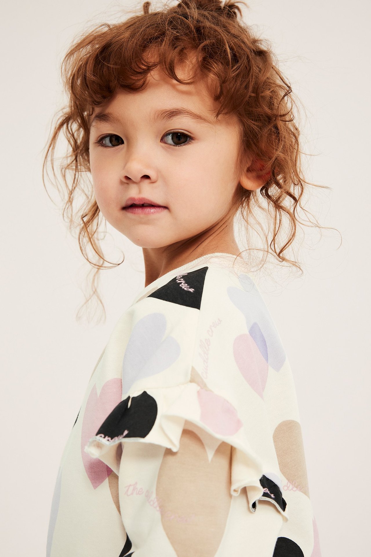 Widok z przodu: Dziecko nosi kremową bawełnianą bluzę z kolorowym wzorem serc i falbankami na rękawach.