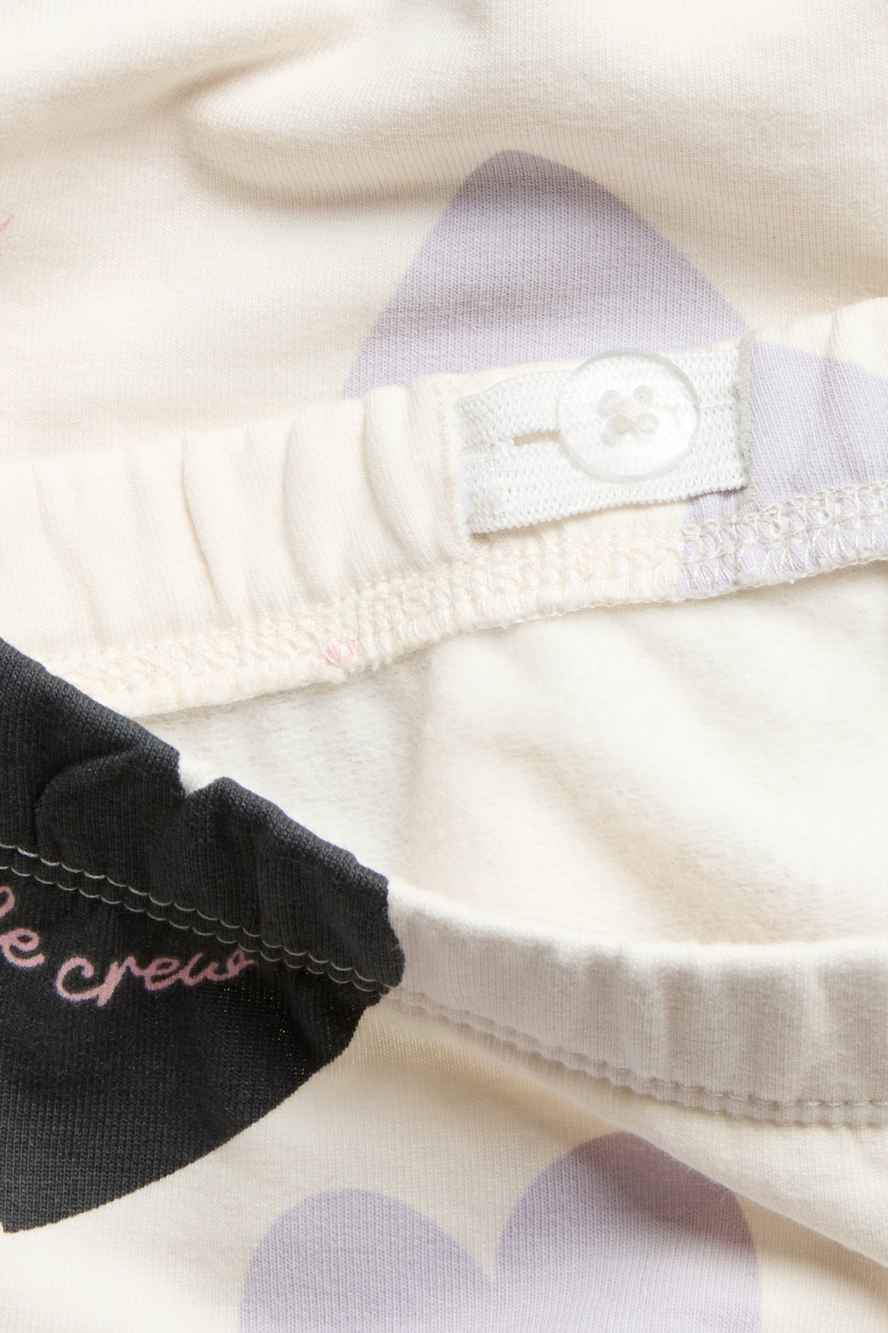 Zbliżenie na dziecięce prążkowane legginsy ecru z fioletowym wzorem serc i regulowanym guzikiem w pasie.