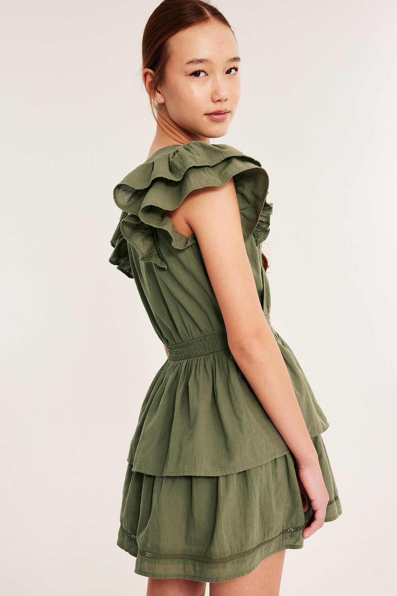 Widok z boku dziecka w ciemnozielonej bawełnianej sukience: falbany, marszczona talia, warstwowa spódnica.