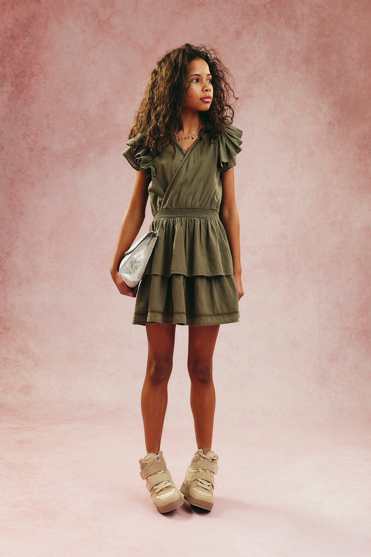 Dziecko w ciemnozielonej sukience z bawełnianymi falbanami i marszczoną talią, trzyma srebrną kopertówkę, beżowe trampki.