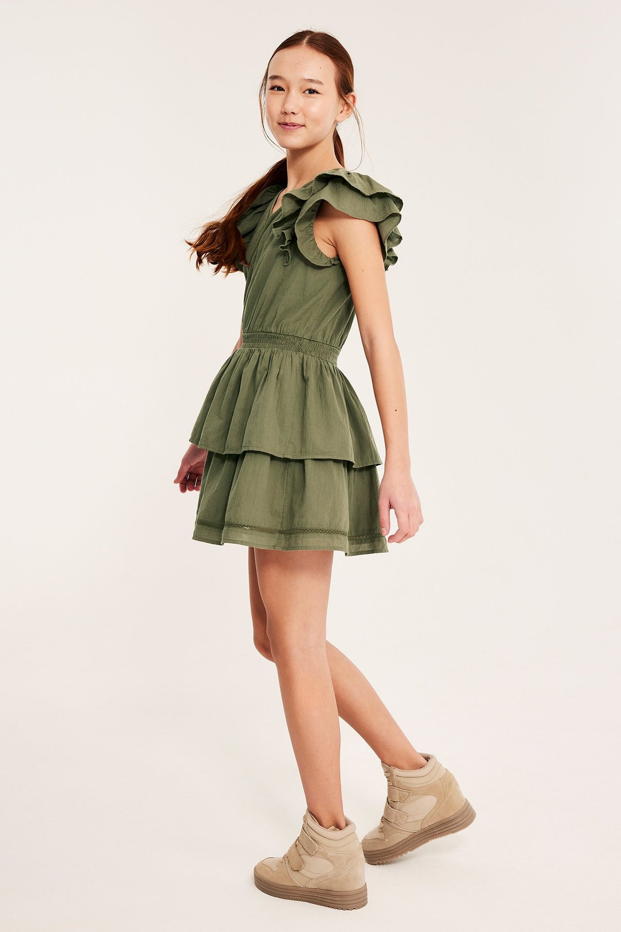 Sideutsikt av et barn i mørkegrønn volang-bomullskjole med smock i midjen, iført beige høye sneakers.