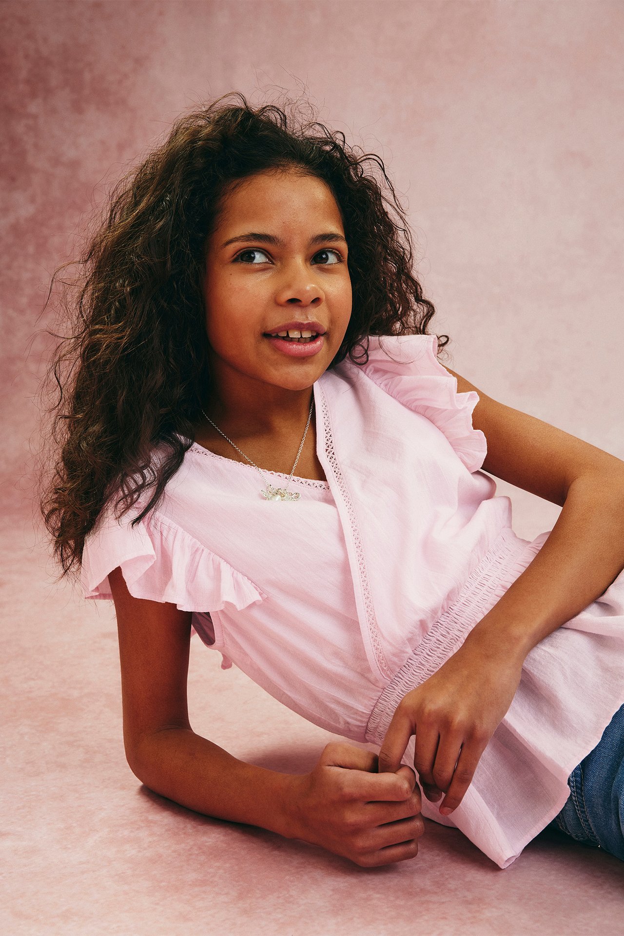 Frontvy: Barn i ljusrosa bomullsvolangblus med spets, smockad midja och silverhalsband.