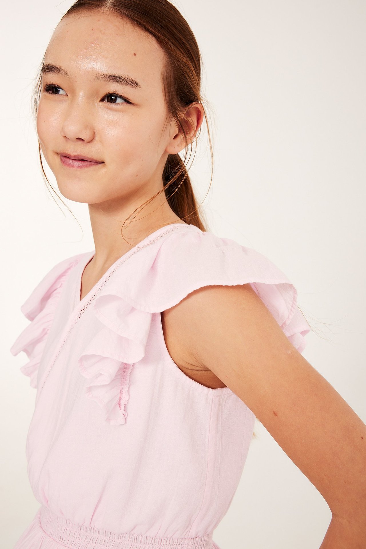 Zbliżenie z boku na dziecko w jasnoróżowej bawełnianej bluzce z falbanami, koronką i ściągaczem w talii.