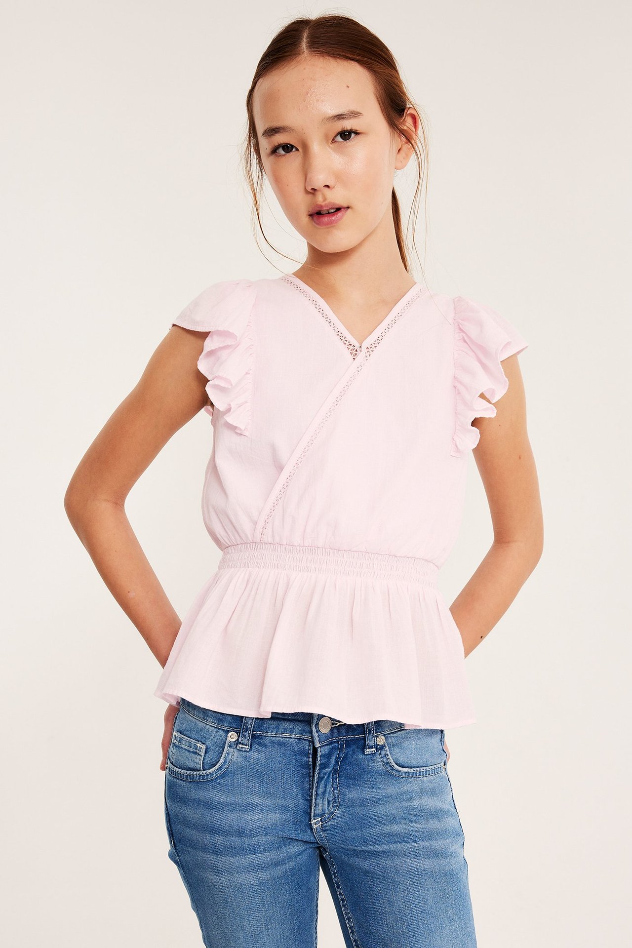 Lapsi edestä: vaaleanpunainen puuvillapusero röyhelöhihoilla, pitsikoristein ja siniset farkut.