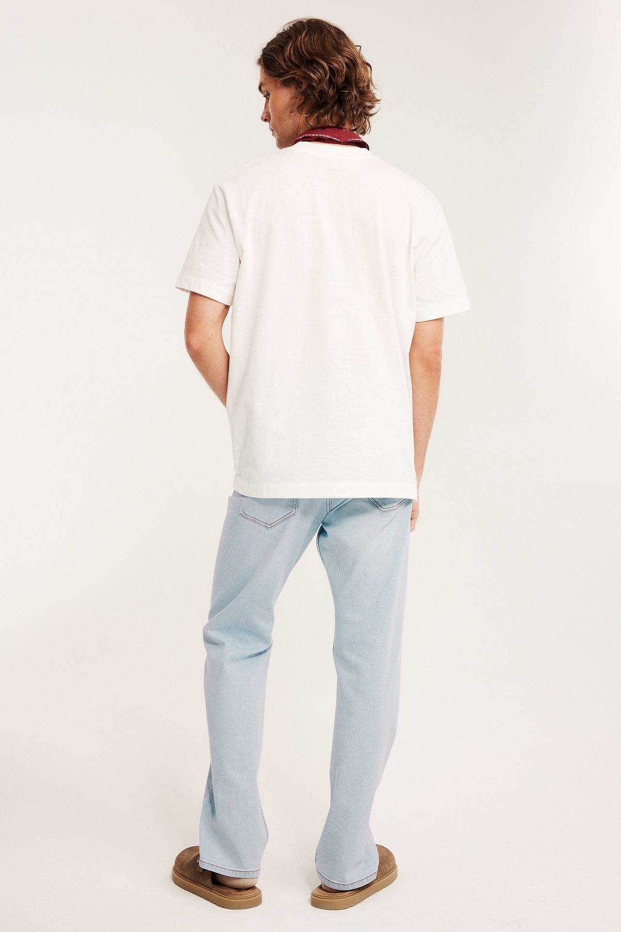 Takaapäin, mies yllään vaaleansiniset regular fit -farkut, valkoinen t-paita, punainen huivi ja ruskeat tohvelit.