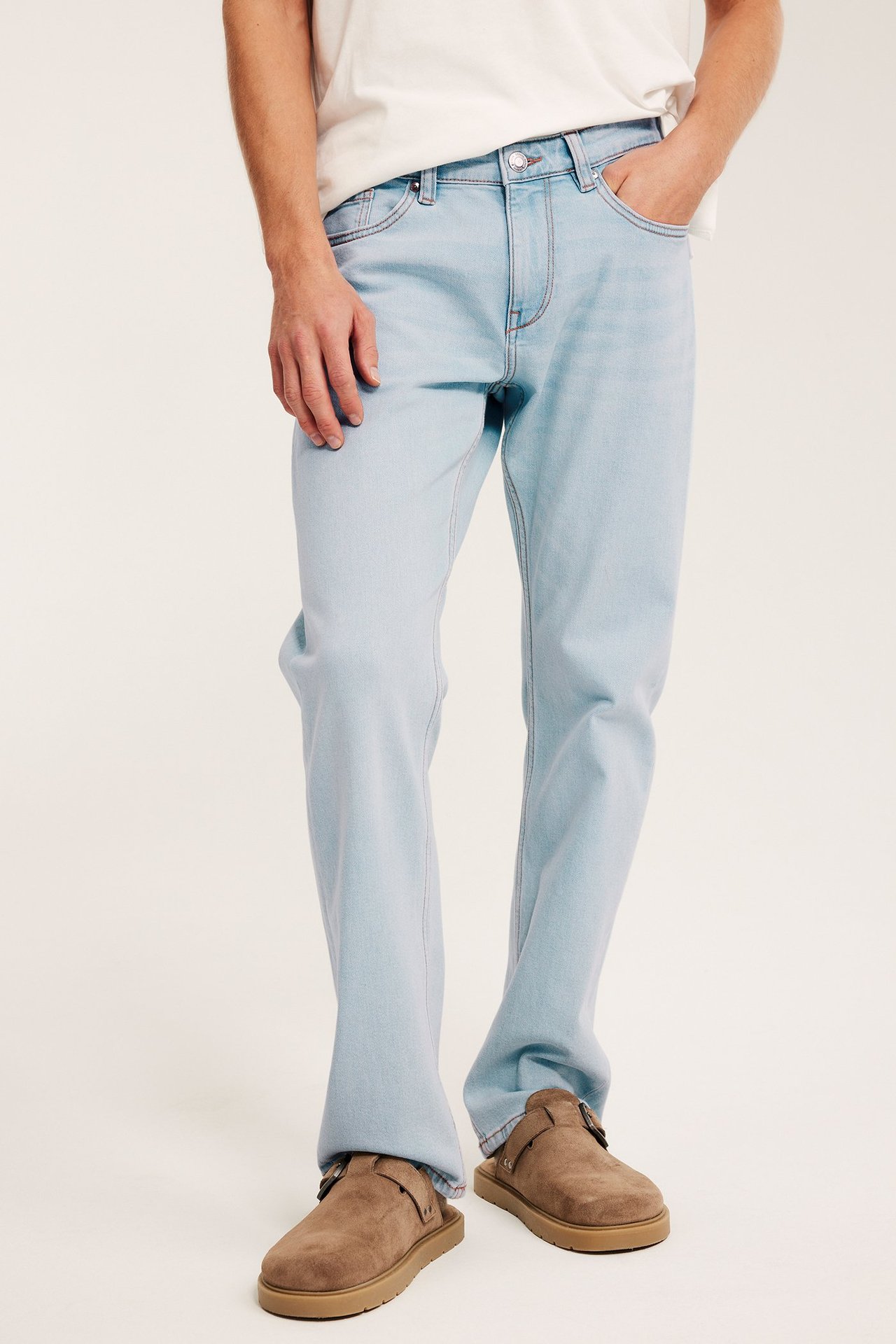 Edestäpäin mies yllään vaaleat regular fit farkut, valkoinen t-paita ja ruskeat solkikengät.