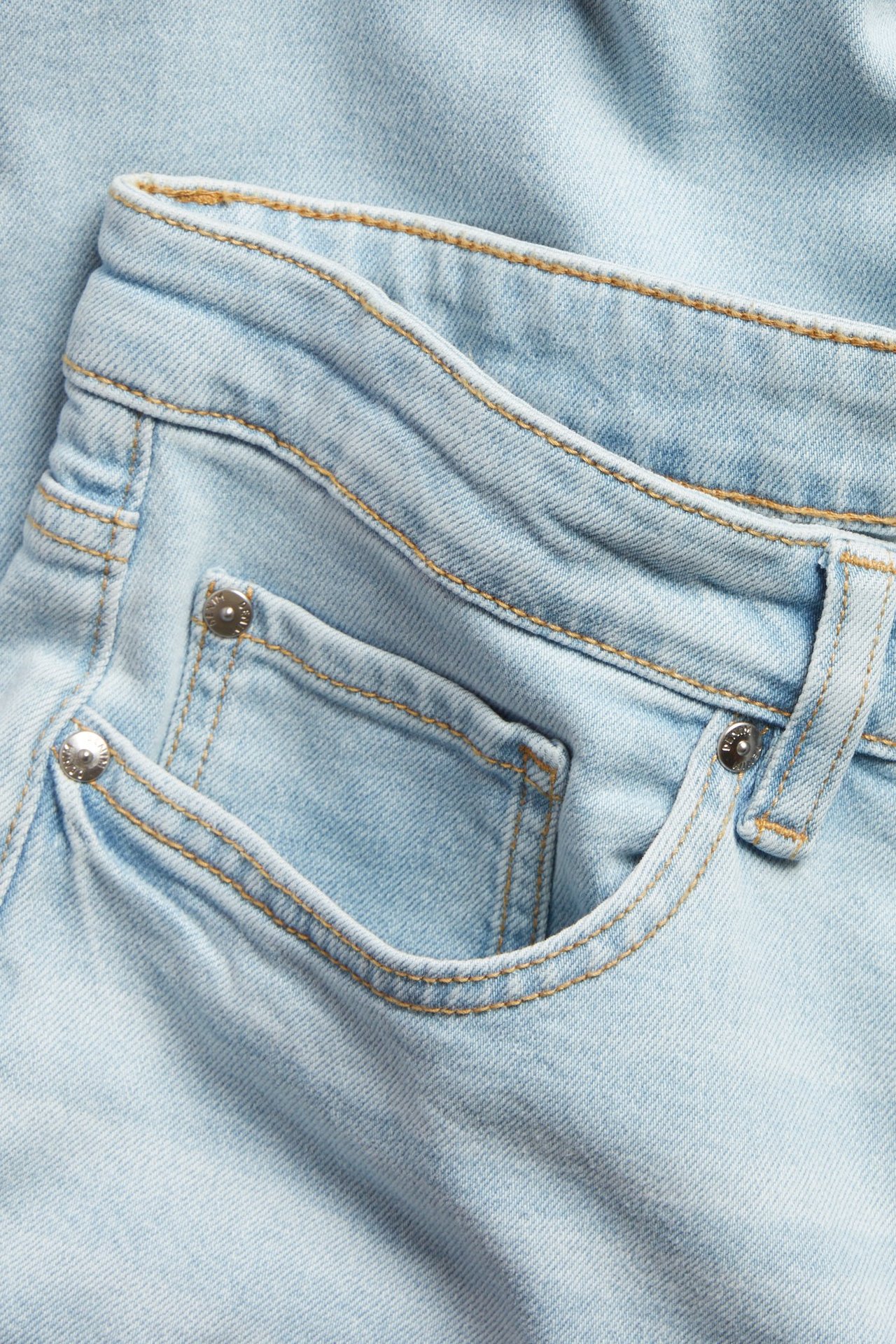 Lähikuva edestä vaaleansinisistä regular fit -farkuista oransseilla tikkauksilla ja metalliniiteillä.