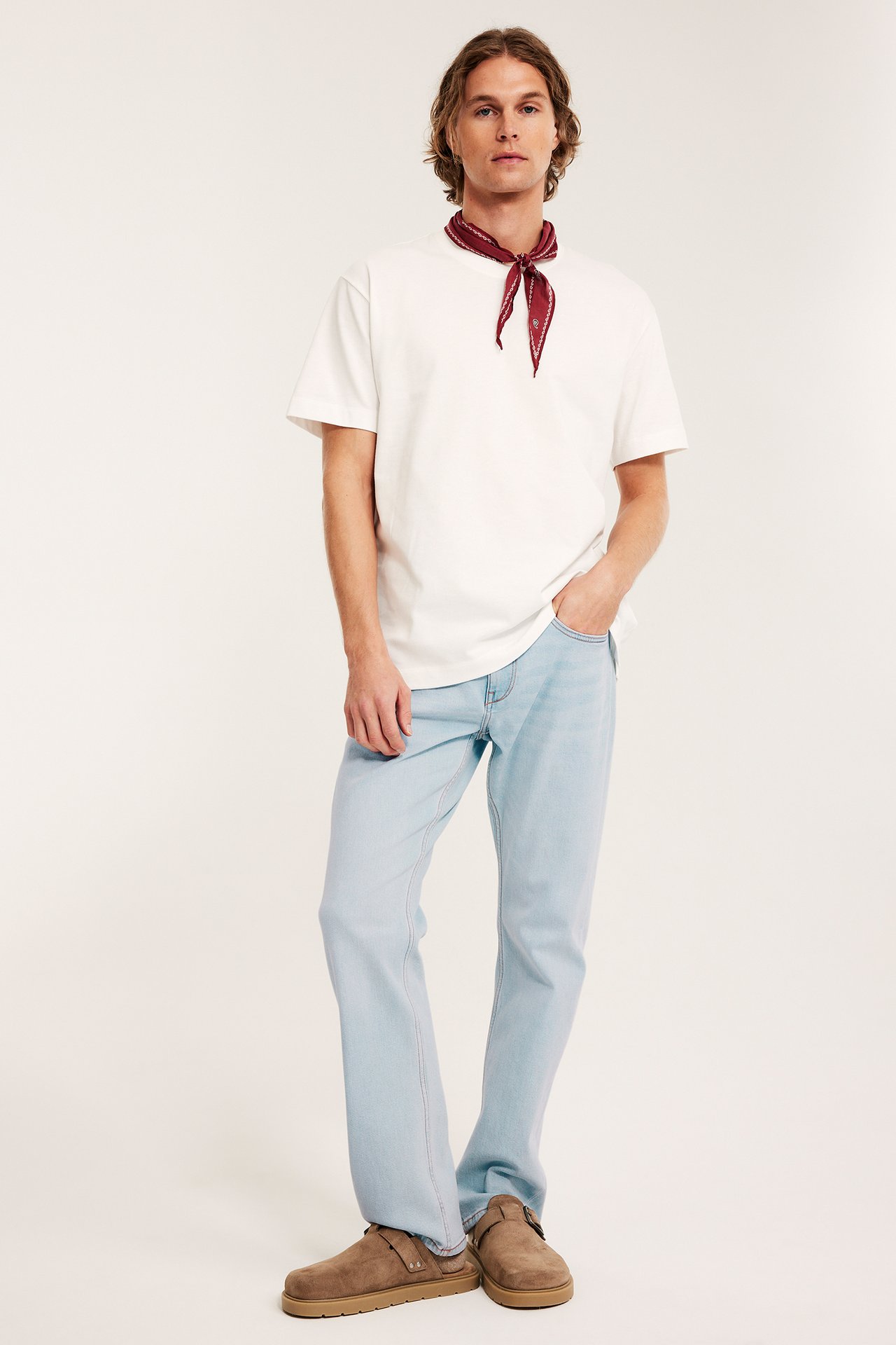 Edestäpäin mies vaaleansinisissä farkuissa, valkoisessa t-paidassa, punaisessa bandanassa ja ruskeissa tohveleissa.