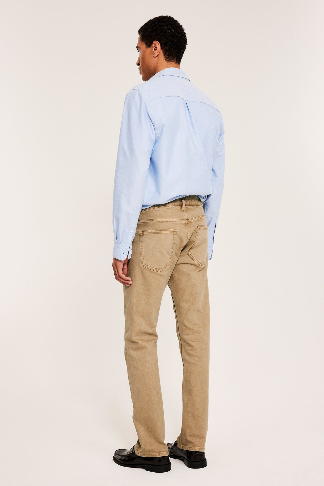 En man i beige regular twill-jeans, ljusblå skjorta och svarta loafers, sedd bakifrån.