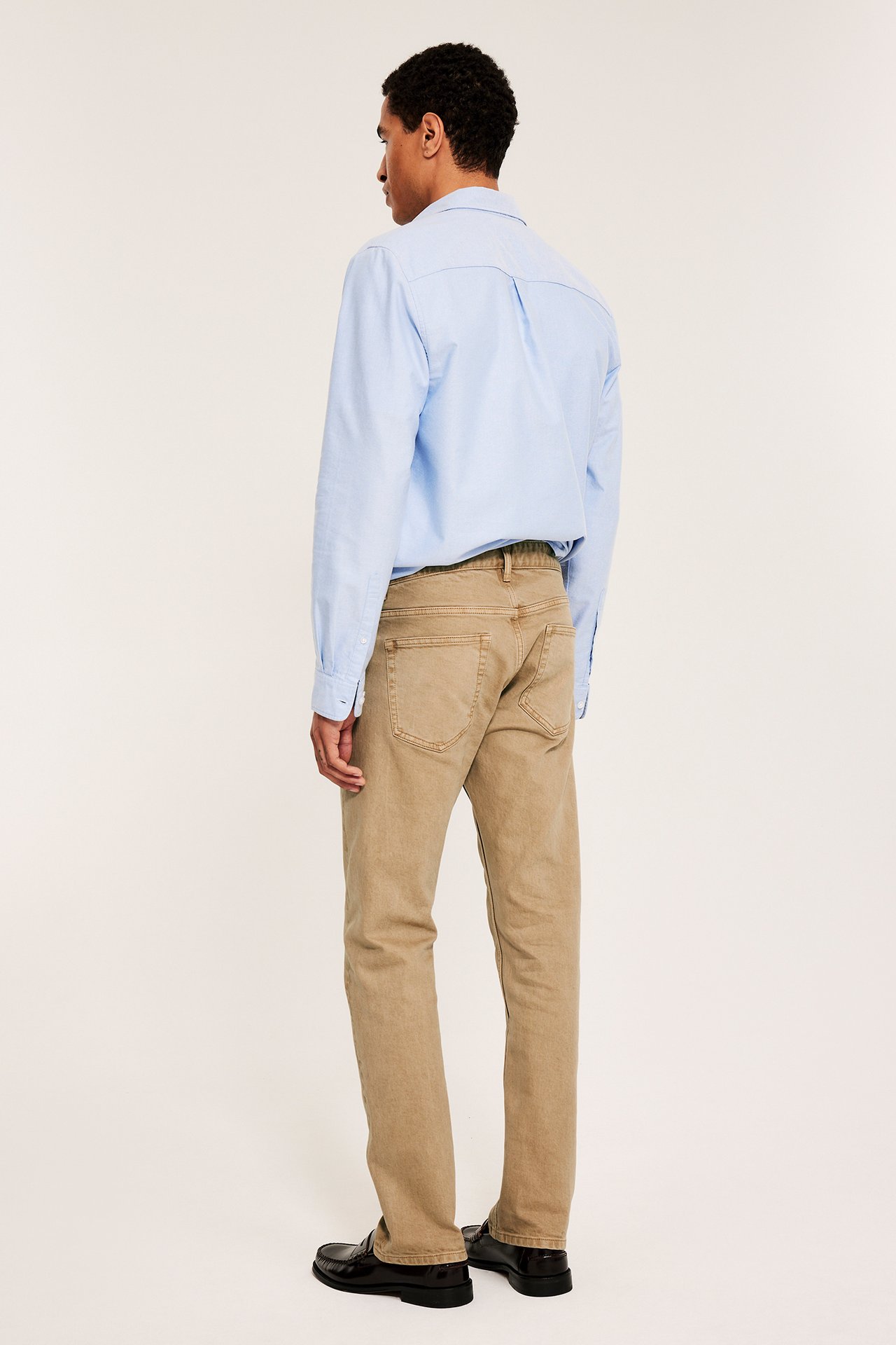 En man i beige regular twill-jeans, ljusblå skjorta och svarta loafers, sedd bakifrån.