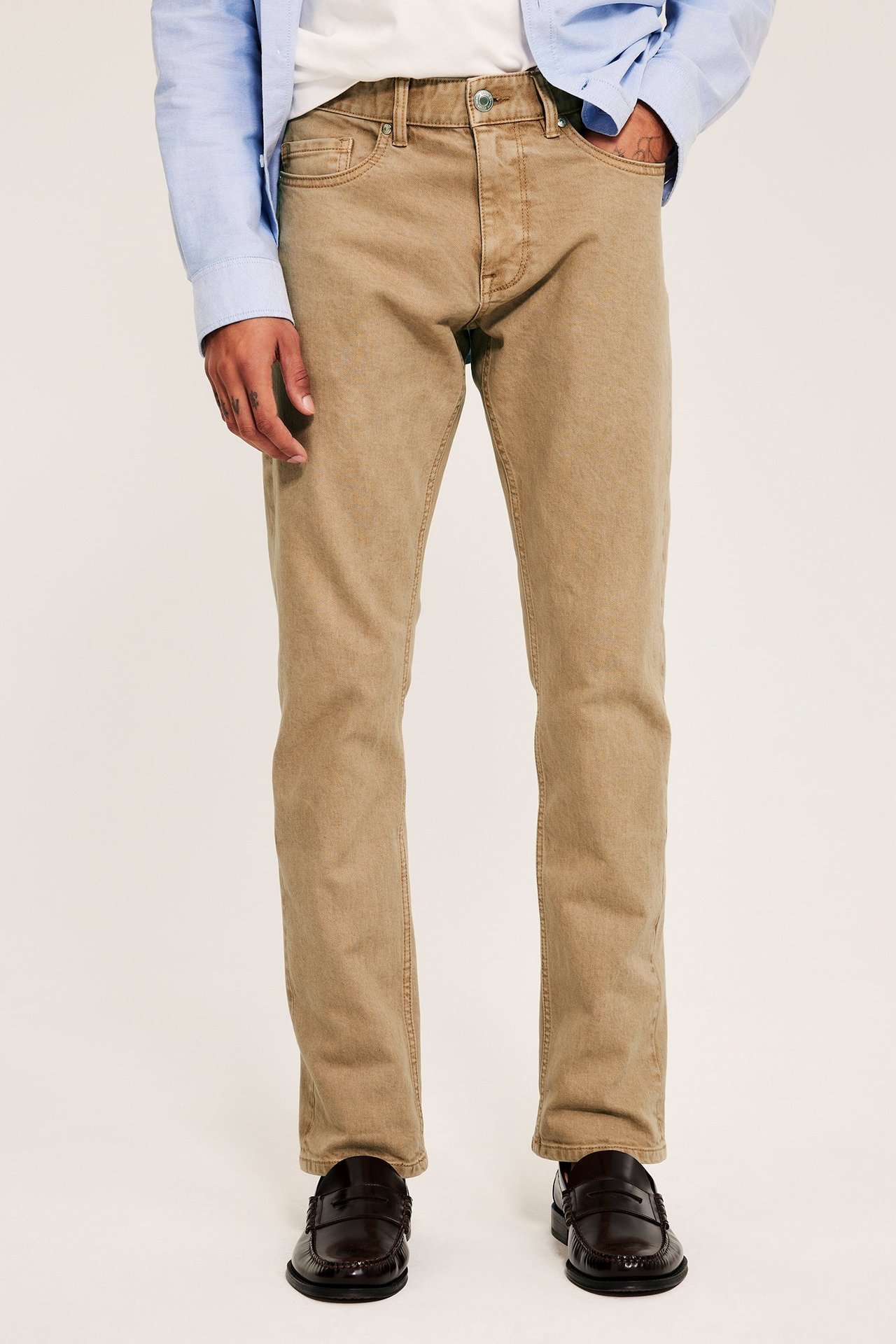 Front view: Man i beiga raka twilljeans, vit t-shirt, ljusblå skjorta och bruna loafers.