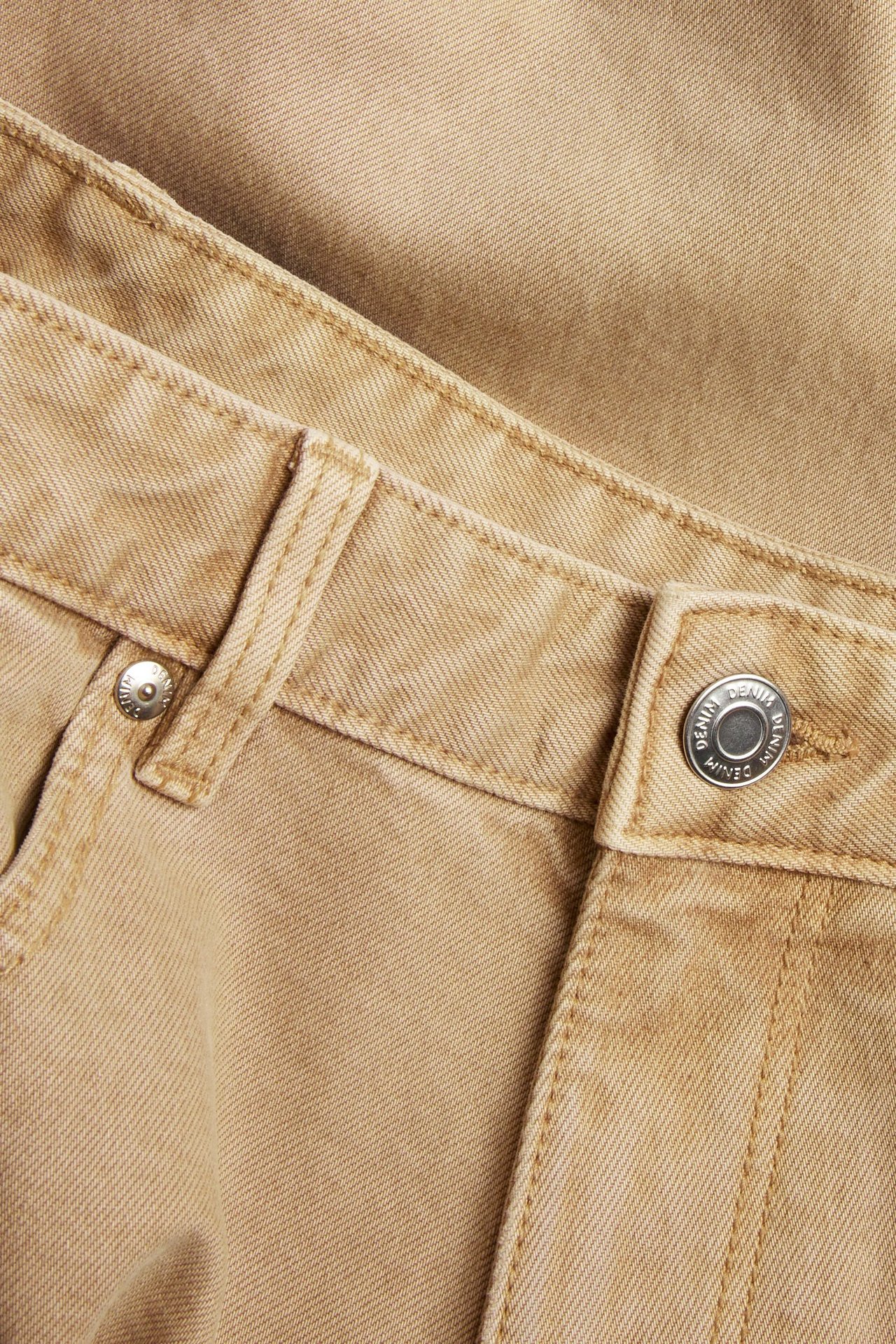Nærbilde forfra av beige regular twilljeans, som viser en sølvknapp og beltehemper.