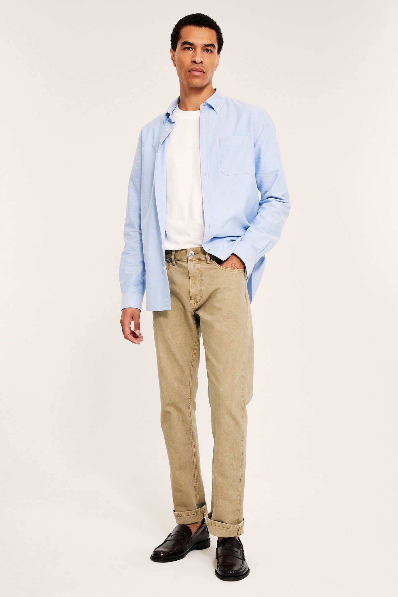 Edestä näkymä miehestä yllään beiget suoralahkeiset twillfarkut, vaaleansininen paita valkoisen t-paidan päällä ja ruskeat loaferit.