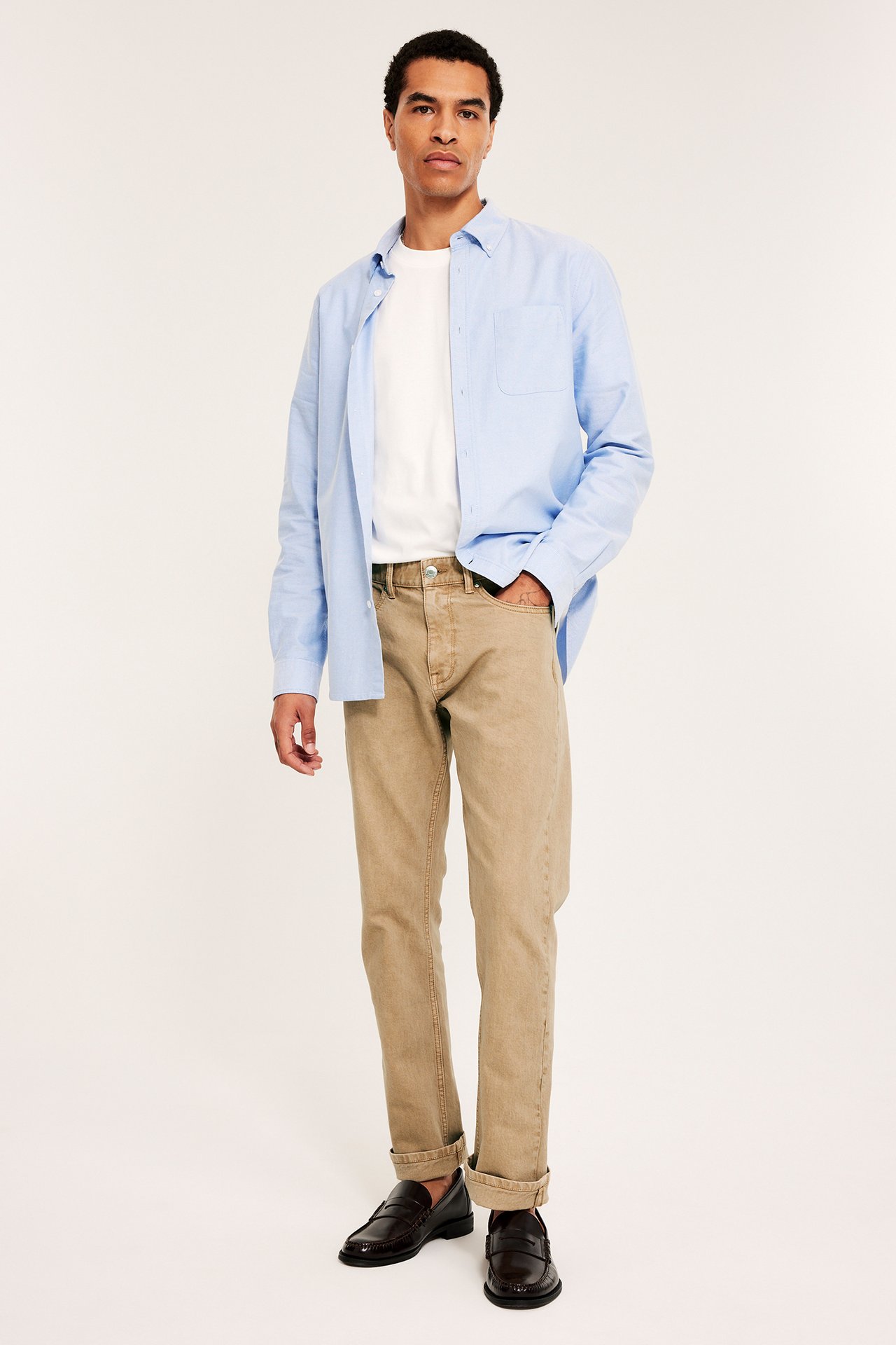 Frontvisning av mann i beige regular twilljeans, hvit t-skjorte, åpen lyseblå skjorte og mørkebrune loafers.