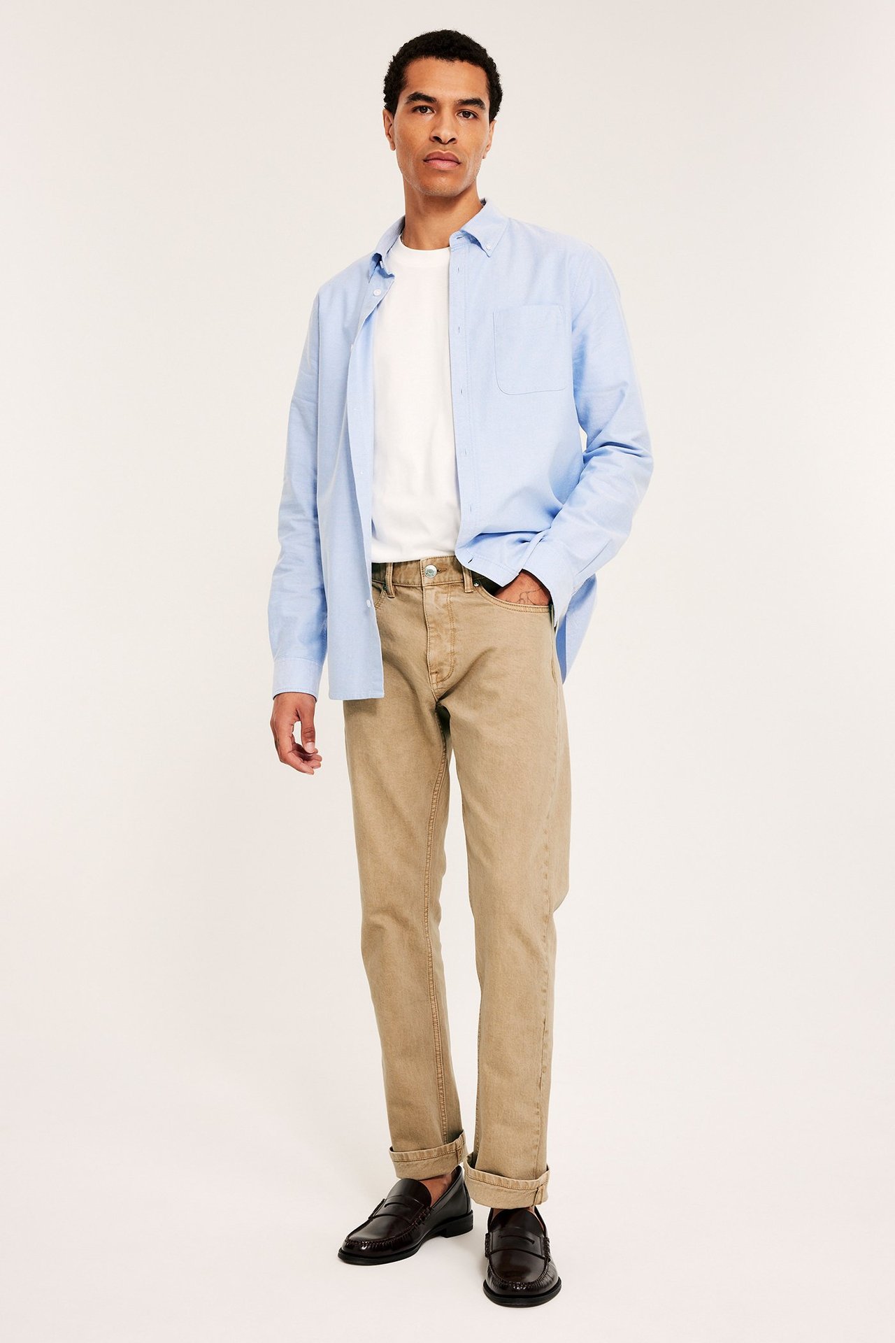 Framifrån en man i beiga raka twilljeans, en vit t-shirt, en öppen ljusblå skjorta och mörkbruna loafers.