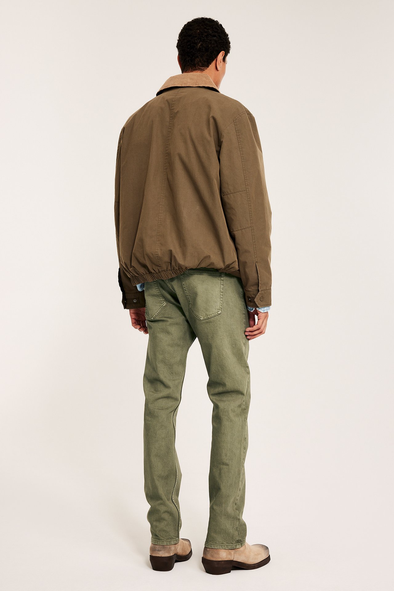 Mies takaa, vihreät suorat twillfarkut, ruskea bomber-takki beige kauluksella, vaaleanruskeat kengät.