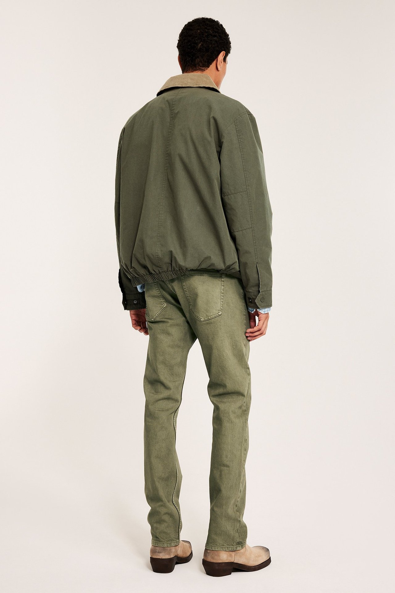 Bakifrån: Man bär gröna regular fit twilljeans, olivgrön jacka över blå skjorta och beiga stövlar.