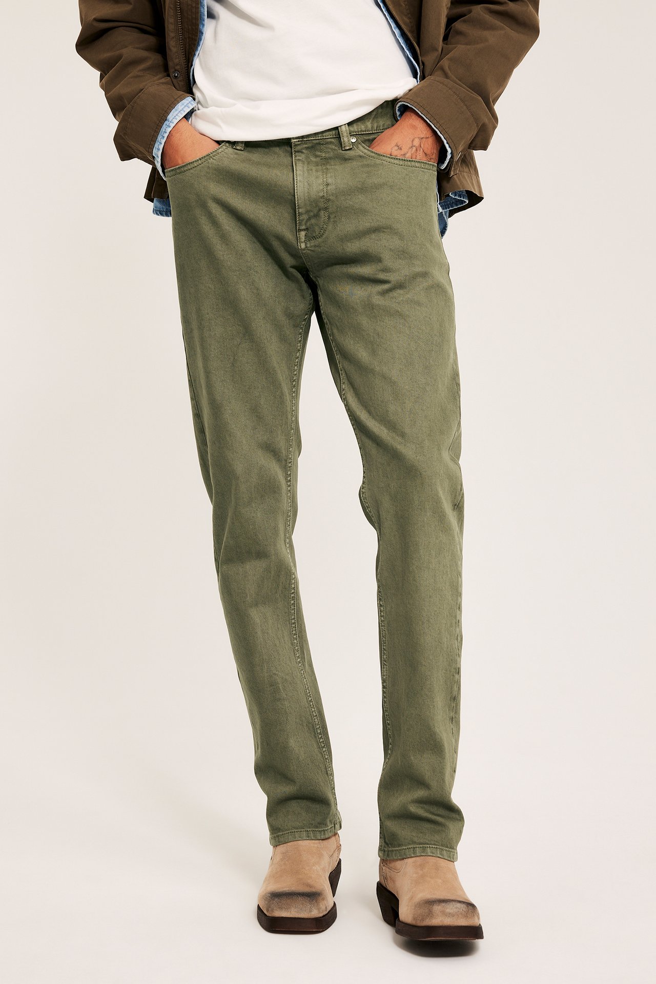 Edestäpäin, mies vihreissä twillfarkuissa, valkoinen t-paita, ruskea takki ja ruskeat nilkkurit.