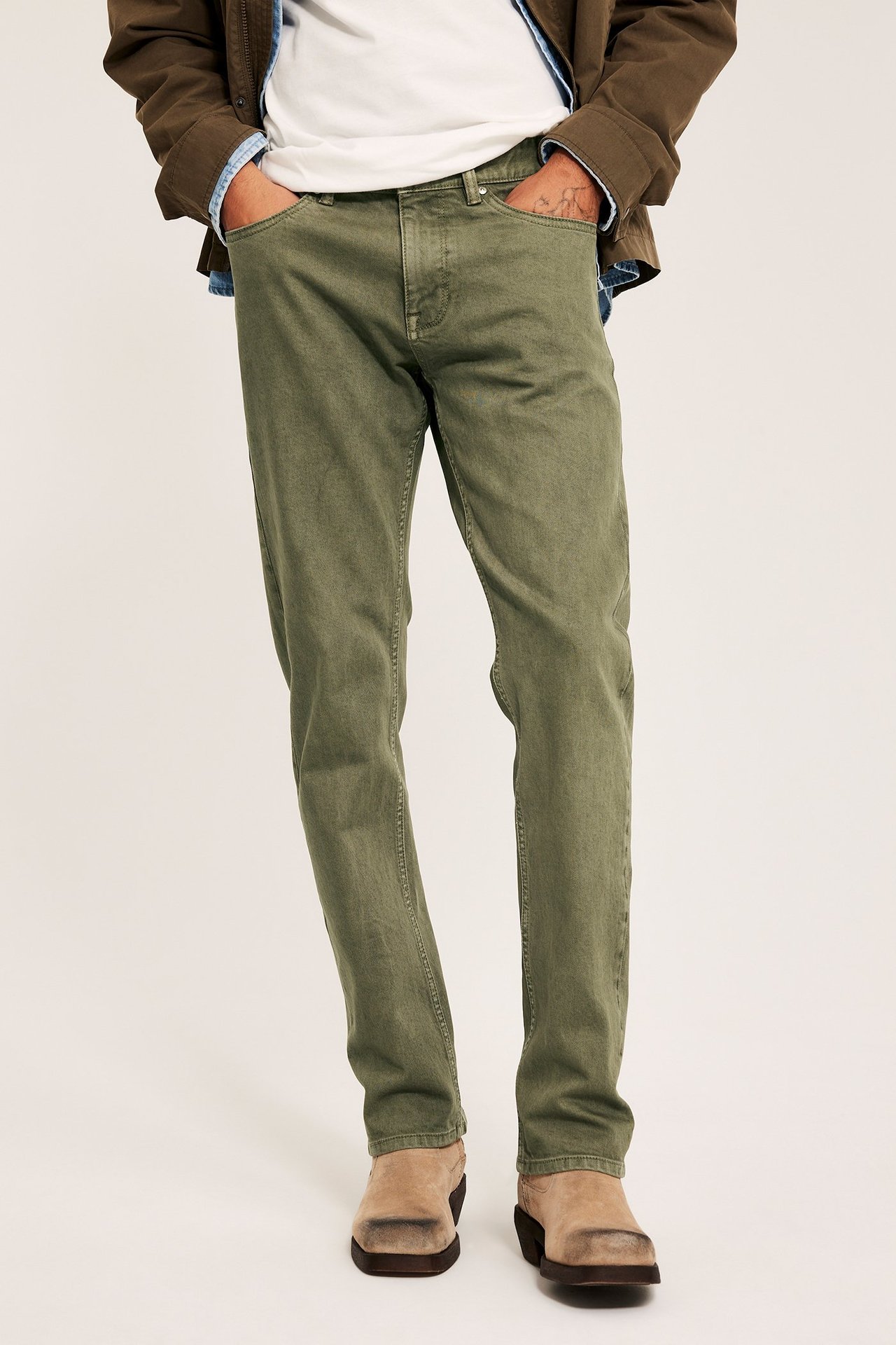 Framifrån, en man i gröna regular twilljeans, vit t-shirt, brun jacka och ljusbruna kängor.