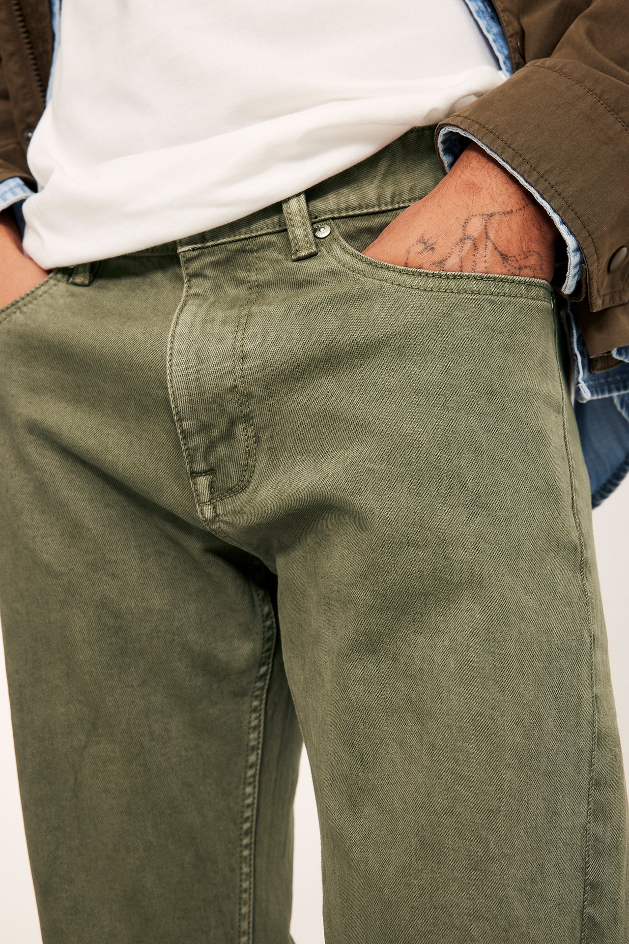 Lähikuva: Mies vihreissä twillifarkuissa, käsi taskussa, valkoinen t-paita ja khakinvärinen takki.