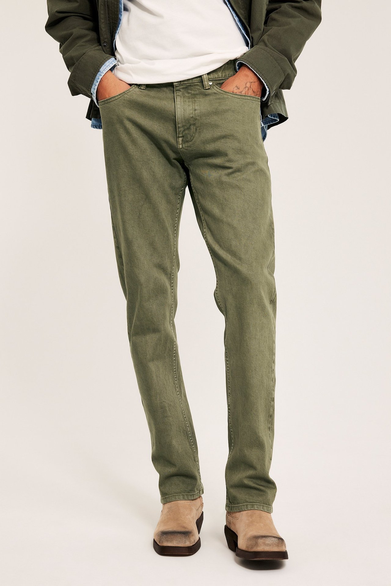 Framifrån: Man i gröna regular fit twilljeans, vit t-shirt, olivgrön jacka och ljusbruna kängor.