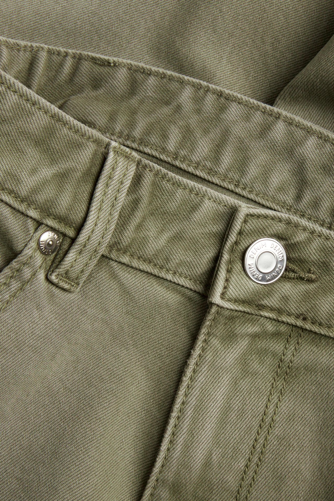 Nærbilde av grønne regular fit twilljeans med linning, beltehemper og en sølvfarget knapp.