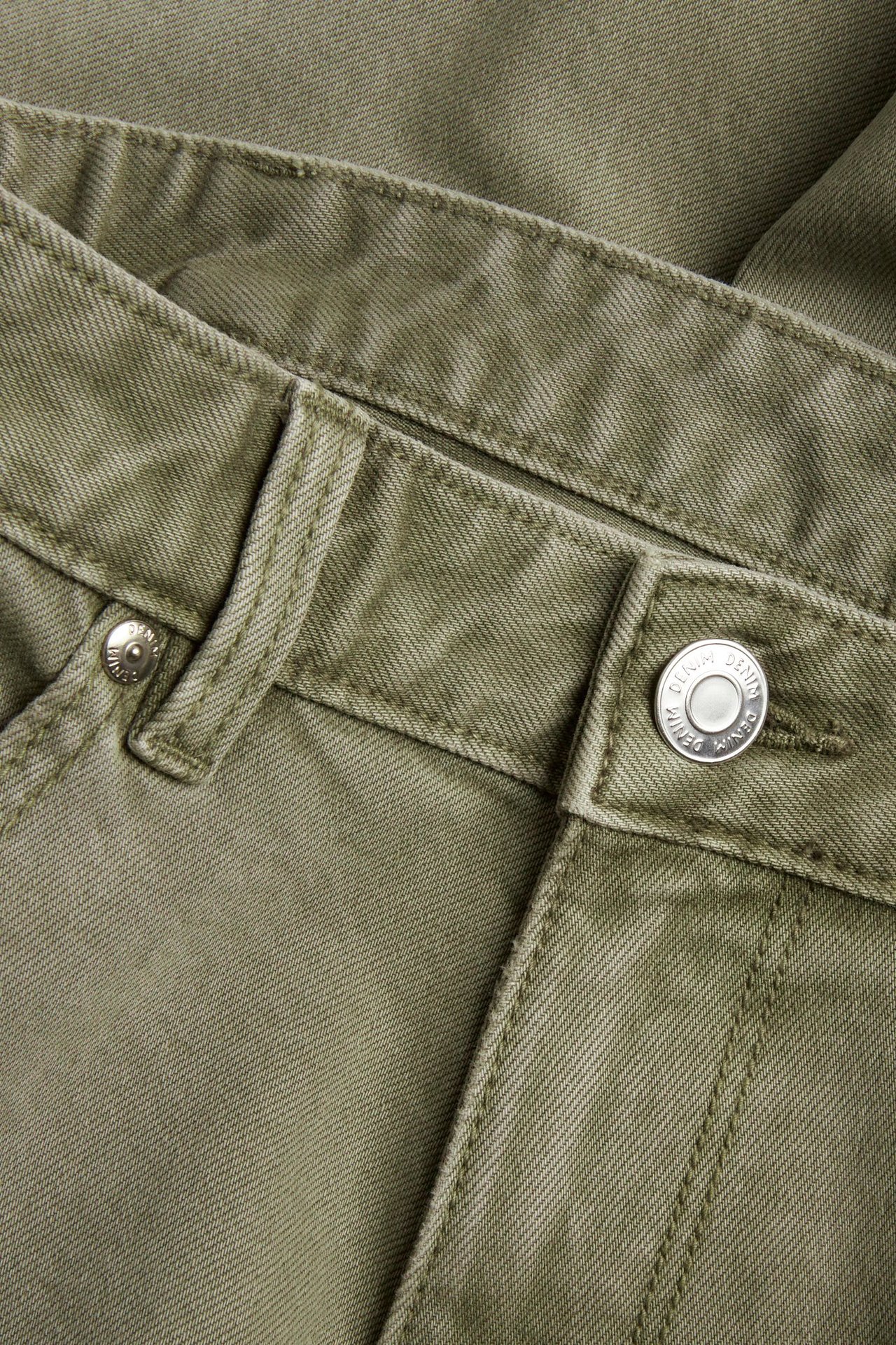 Nærbilde av grønne regular fit twilljeans med linning, beltehemper og en sølvfarget knapp.