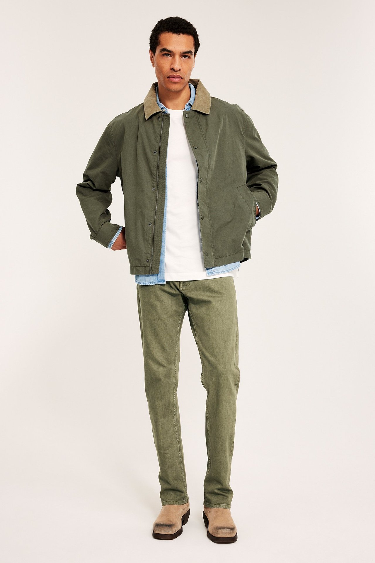 Edestä, mies pukeutuu vihreisiin regular-twill-farkkuihin, valkoiseen t-paitaan, tummanvihreään takkiin ja beigeihin puukenkään.
