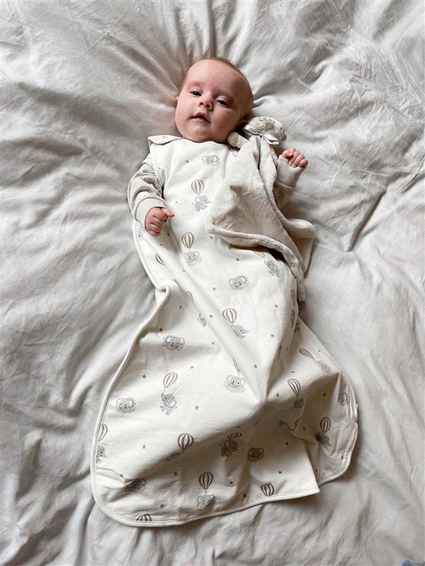 Vauva luonnonvalkoisessa nukkumapussissa norsukuvioinnilla, olkapään neppareilla, puettuna vaalean bodyn päälle.
