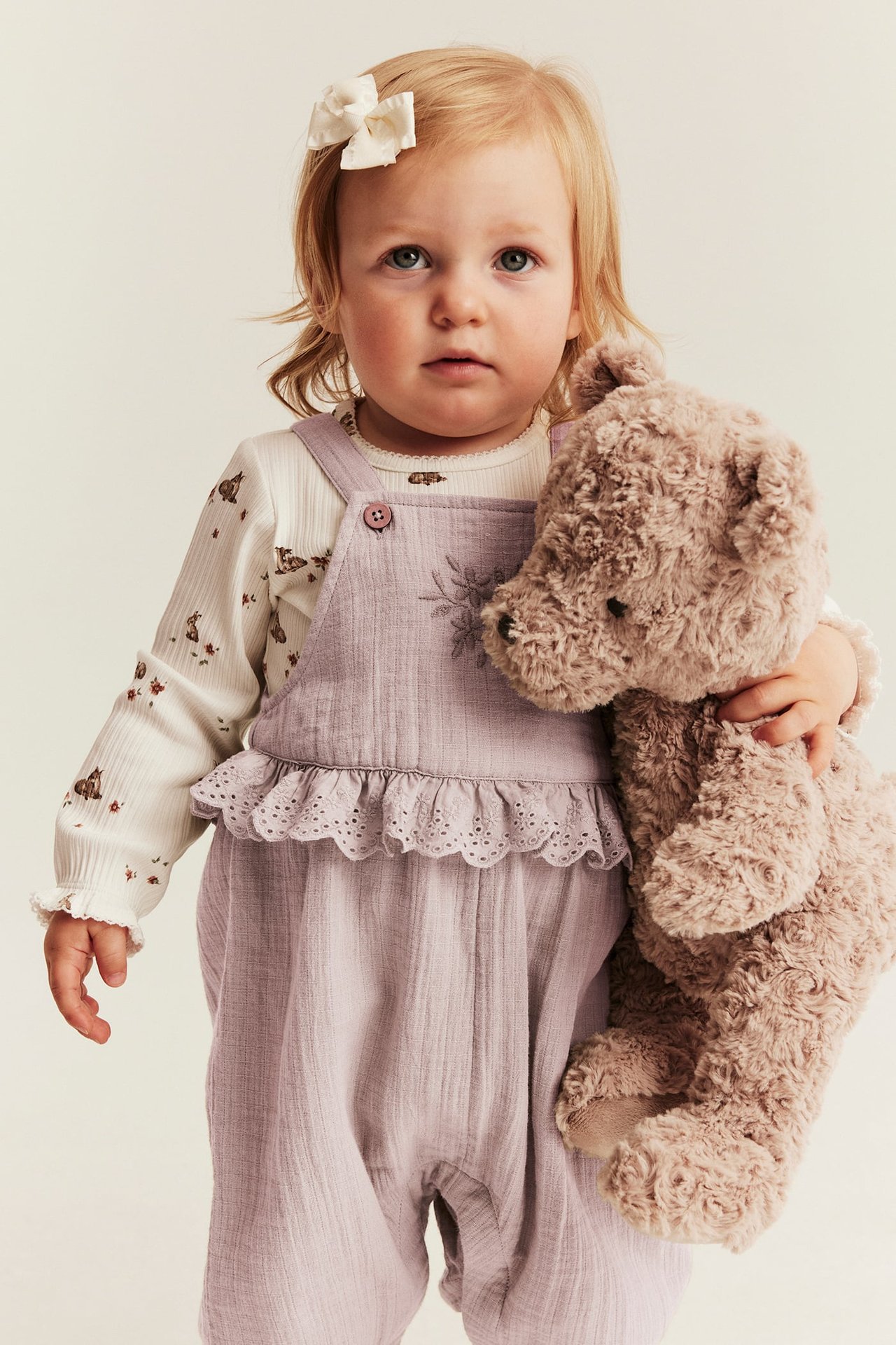 Baby i lyslilla bomullsselebukser med blonderusk og blomsterbroderi, holder en brun teddybjørn.