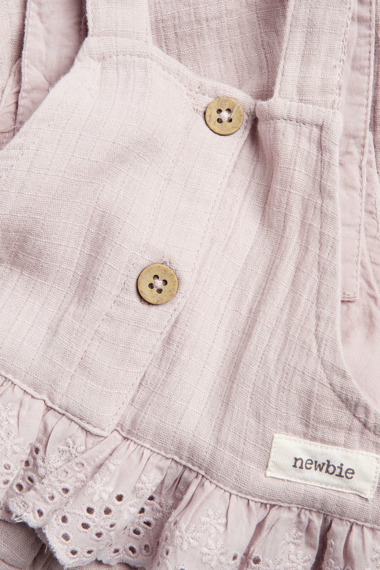 Närbild på ljuslila hängslebyxor i bomull för bebis, med träknappsremmar och spetsvolang.