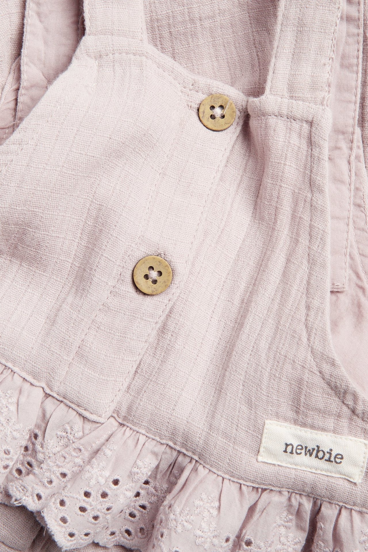 Zbliżenie na jasnoliliowe bawełniane ogrodniczki niemowlęce z drewnianymi guzikami i koronkową falbanką.