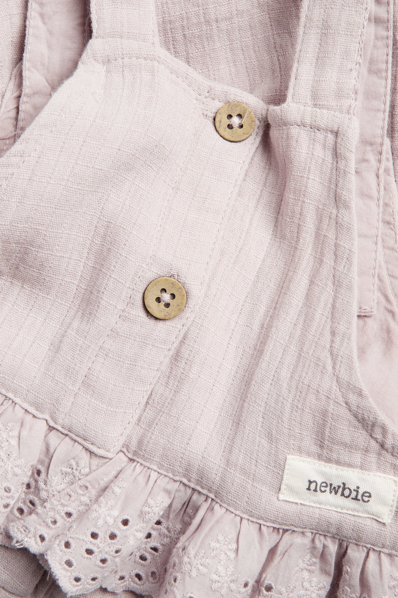 Närbild på ljusrosa muslinoverall för baby med träknappar och broderade volanger.
